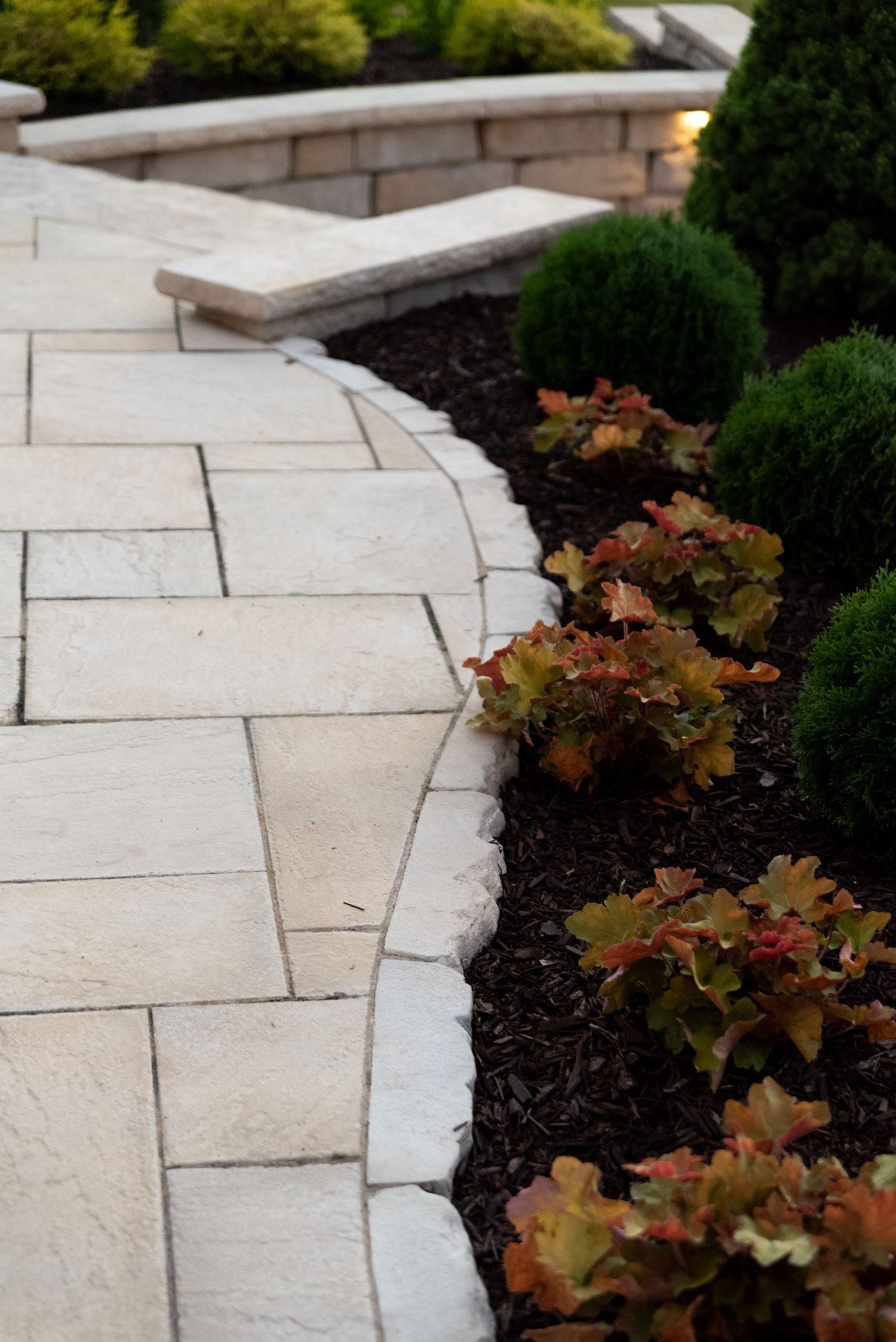 Curved stone walkway bordering a garden bed with green and reddish-brown plants.