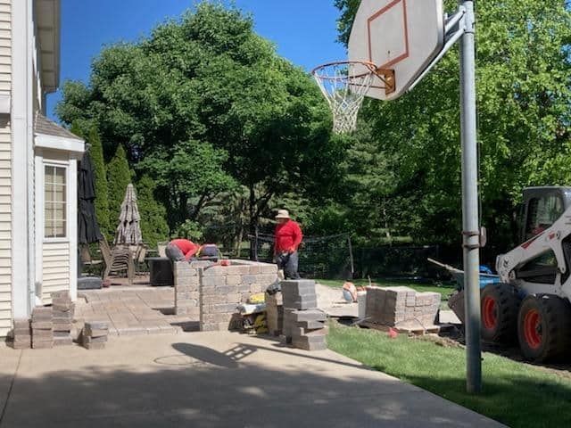 Construction workers build a brick patio near a house with a basketball hoop on a sunny day.