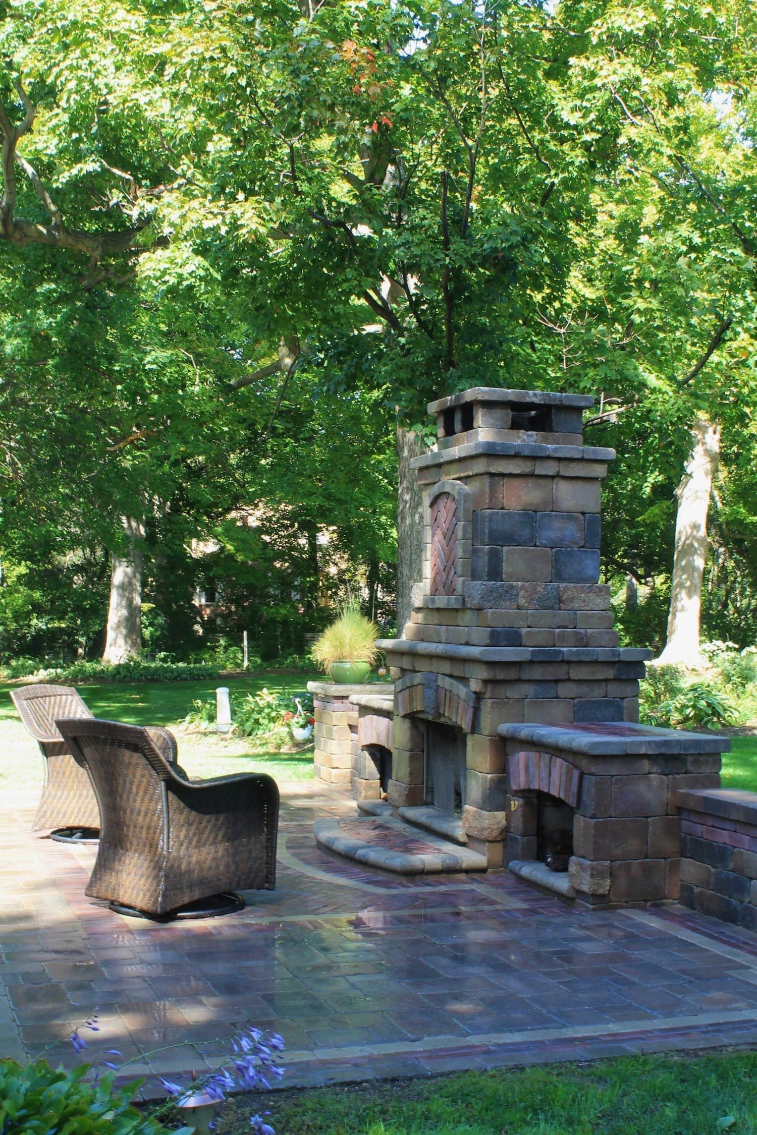 Stone outdoor fireplace with seating on a patio, surrounded by trees and greenery.