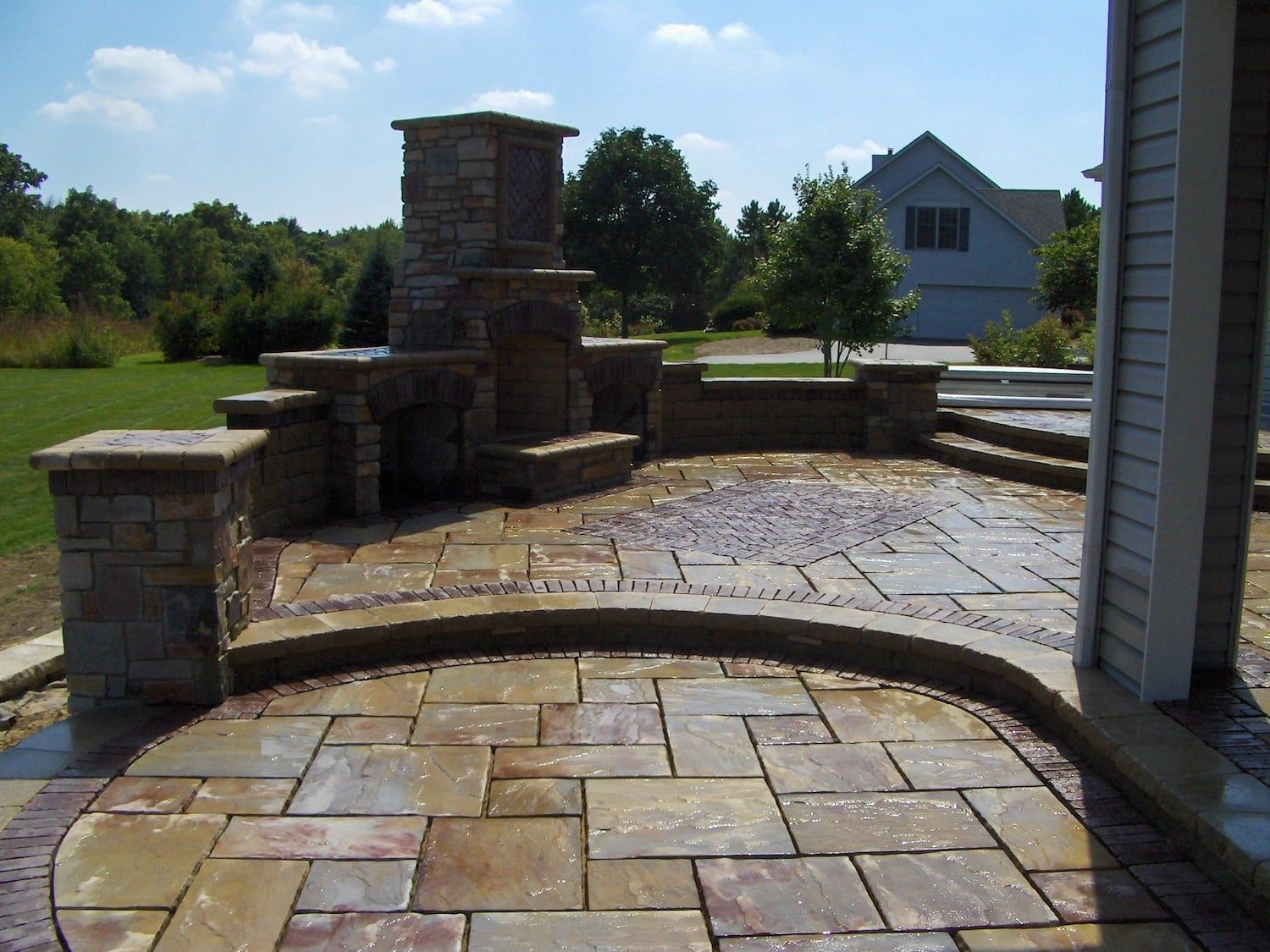 Stone patio with fireplace and pillars next to a house under a blue sky.