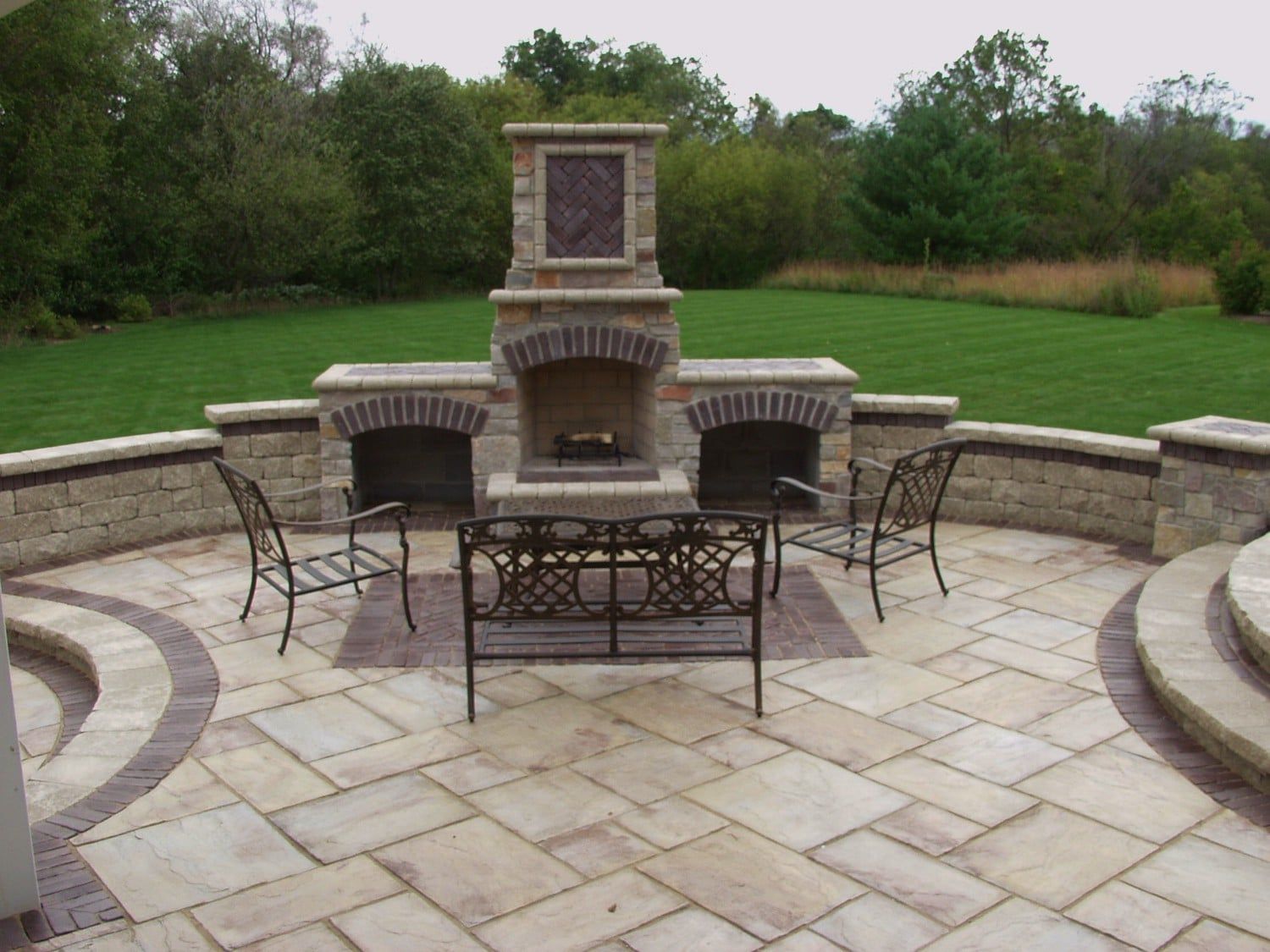 Stone patio with fireplace and seating overlooking a grassy yard.
