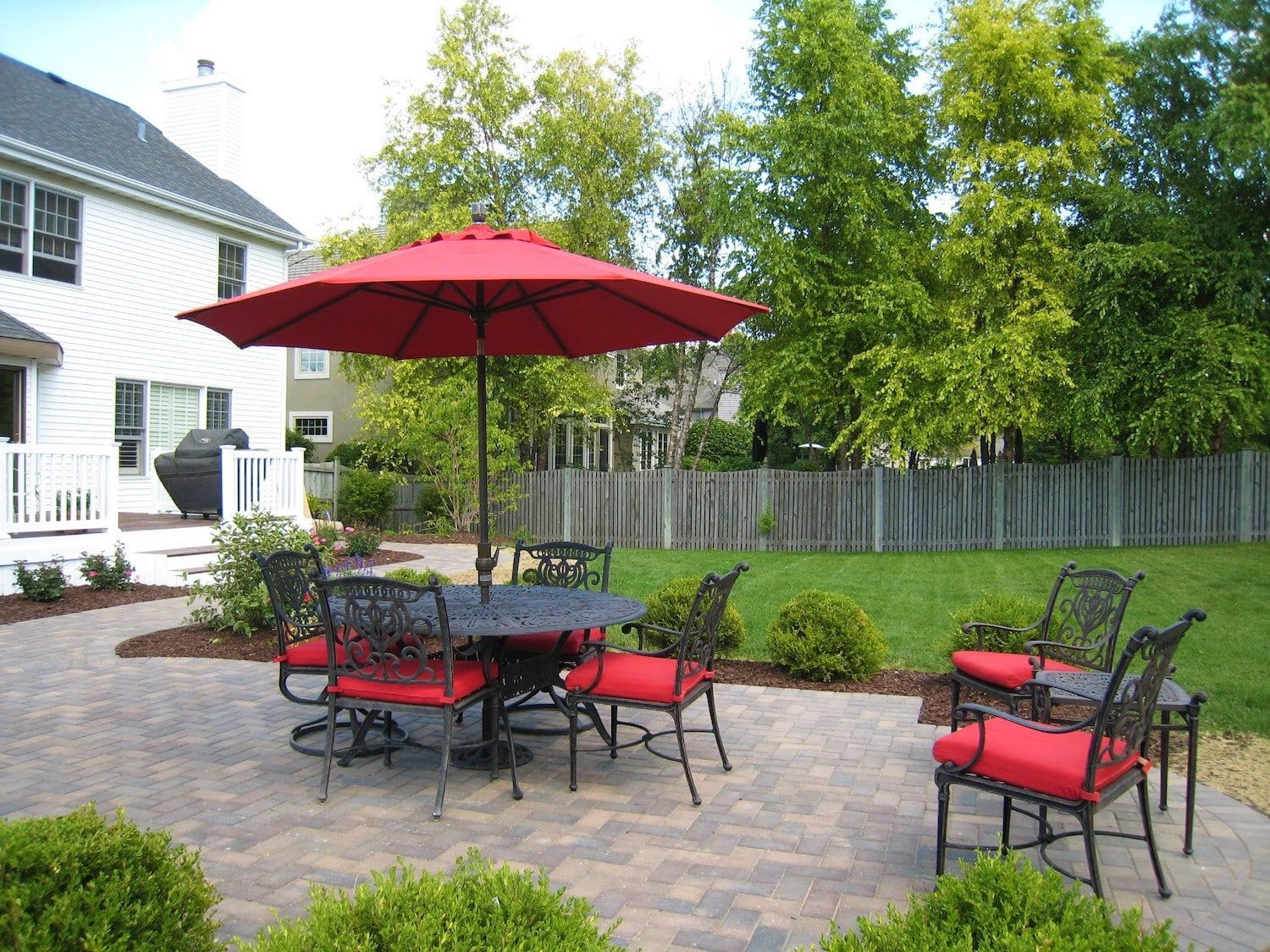Backyard patio with red umbrella, table, and chairs. Green lawn, trees, and house.
