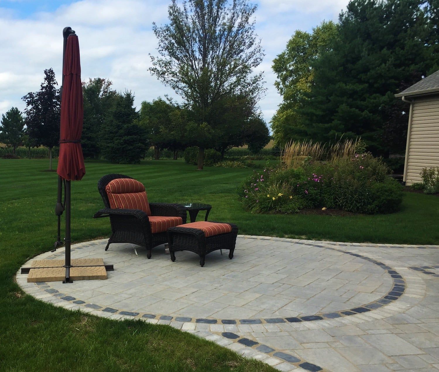 Patio with wicker chair, ottoman, and umbrella on a circular stone surface in a yard.
