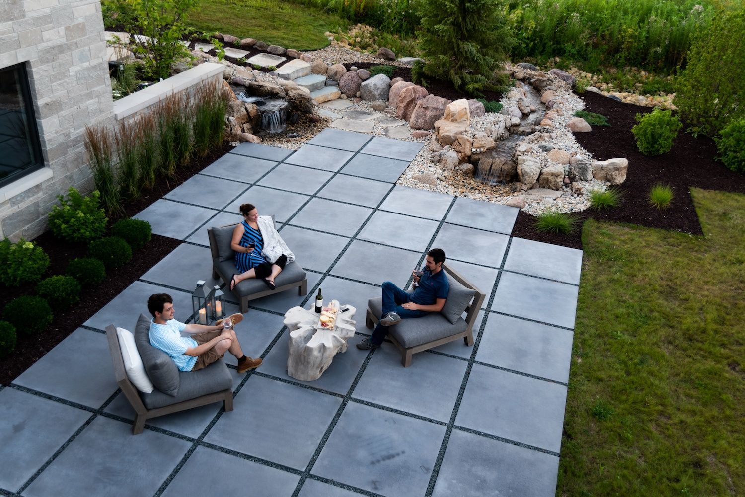 Three people relax on patio furniture on a stone patio; a water feature and lawn are in the background.