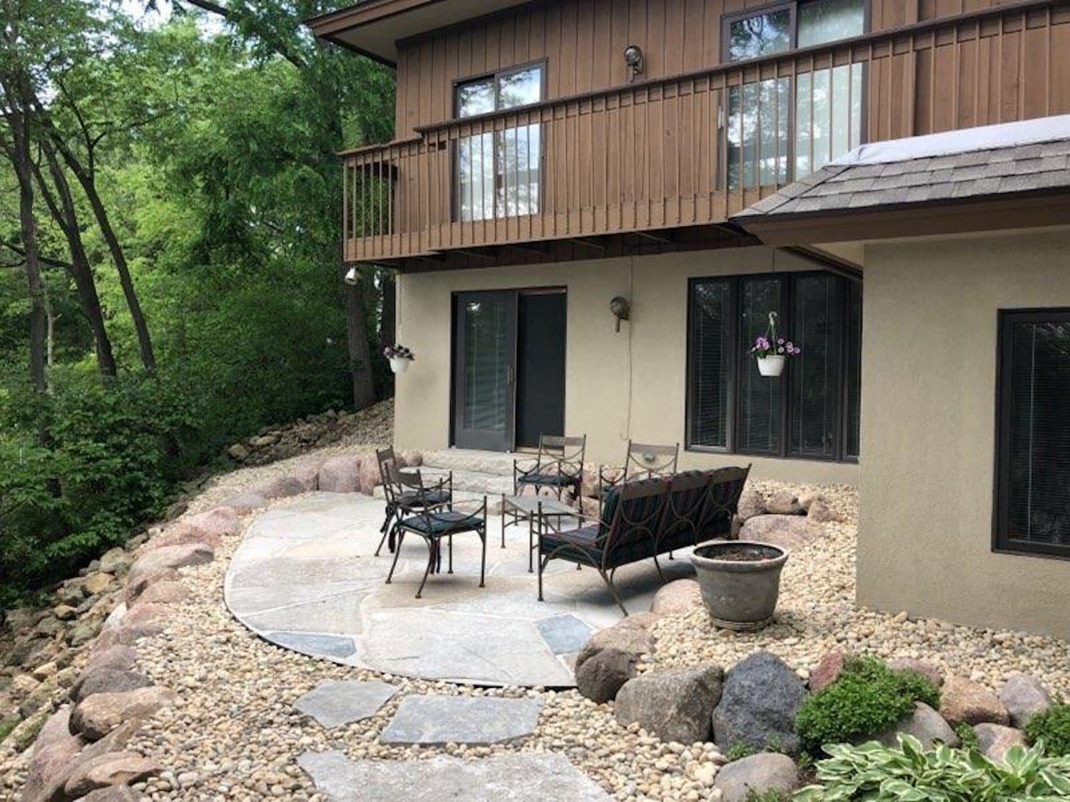 Back patio with stone pavers, seating, and landscaping next to a two-story brown house with a balcony.