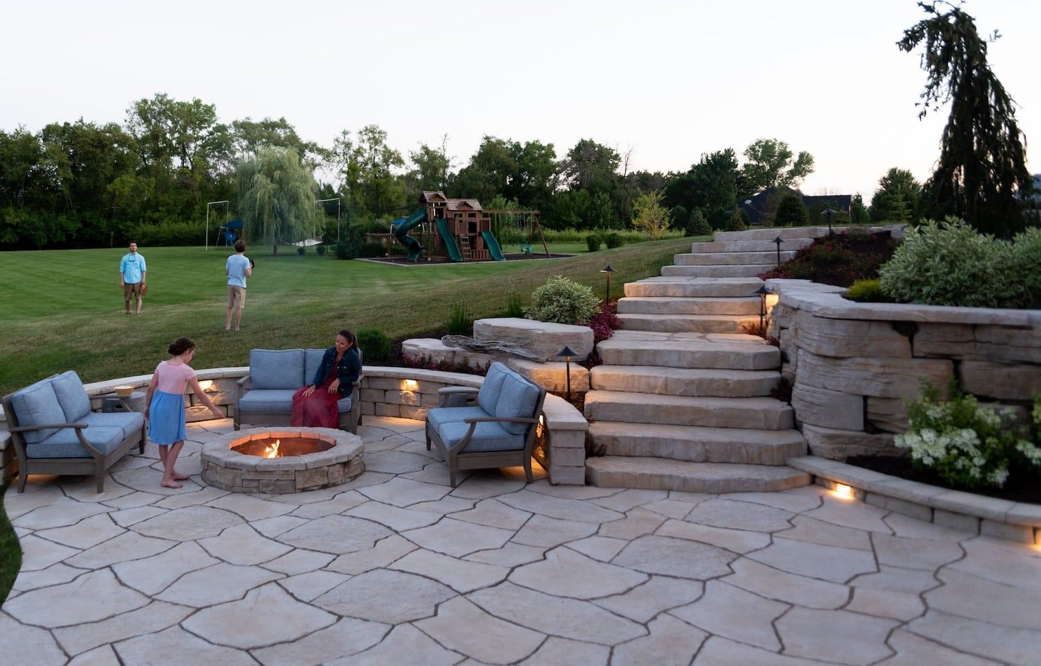 Stone patio with fire pit, seating, and stairs, lit at dusk. People gather in the yard.