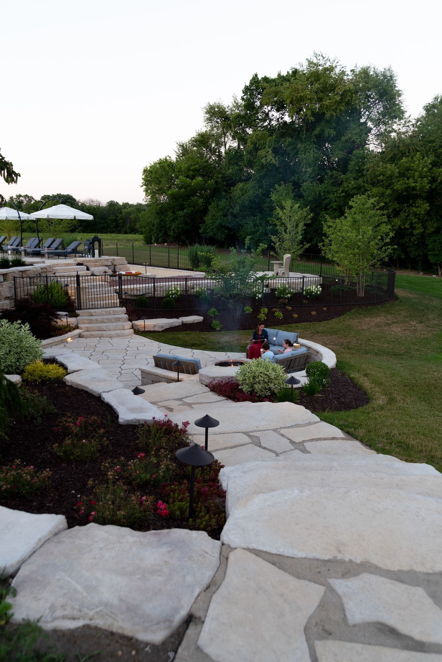 Stone path leads through landscaped yard with lights, toward a pool and trees.