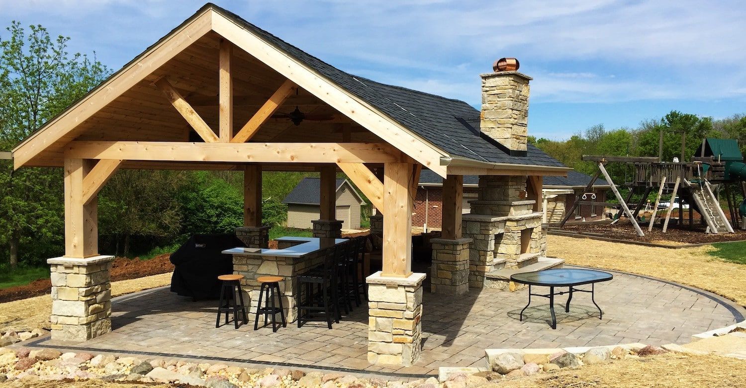 Outdoor kitchen with stone accents, bar, grill, and fireplace under a wooden roof.