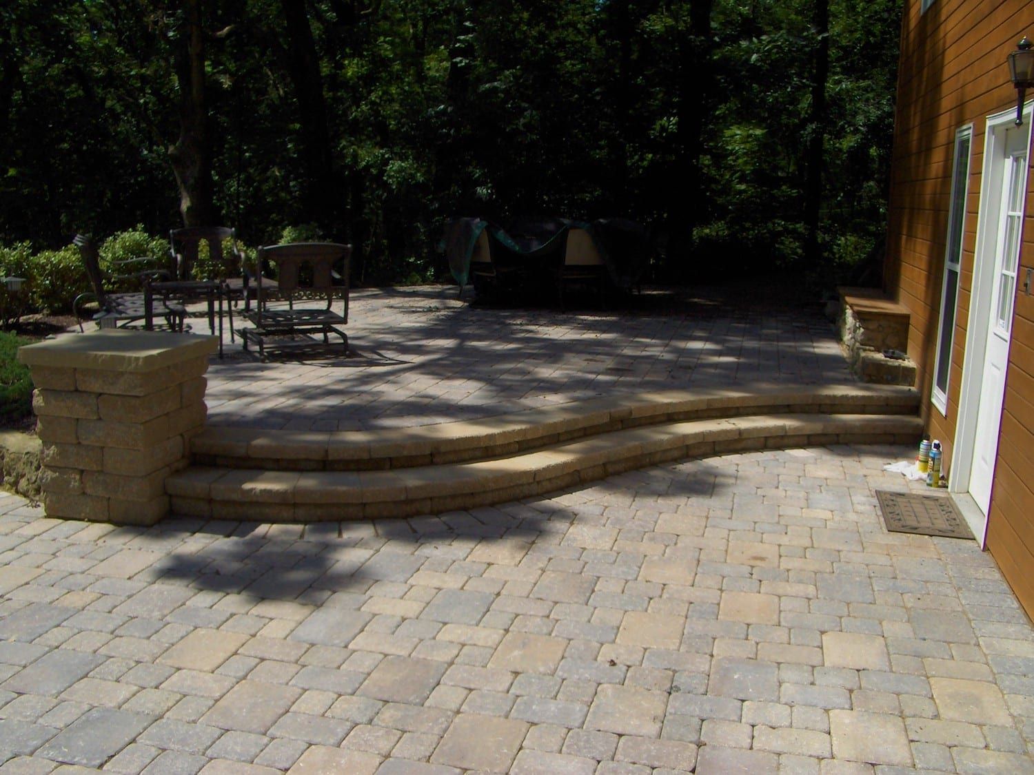 Stone paver patio with steps, column, and door. Outdoor setting with trees and furniture.