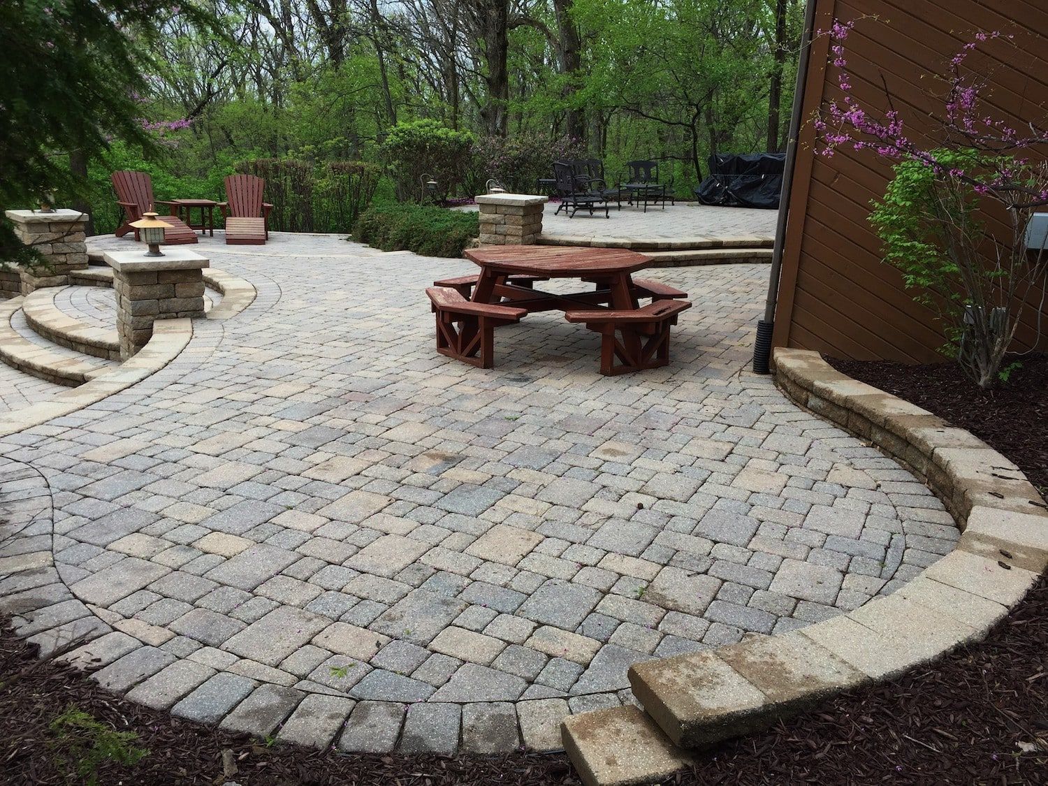 Stone patio with curved border, steps, and picnic table; surrounded by trees and shrubs.