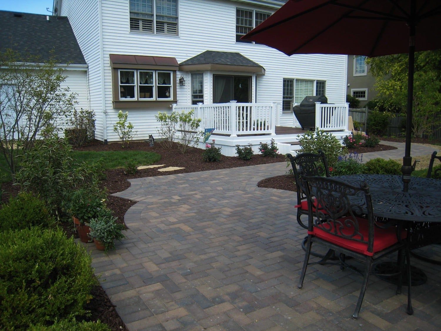 Brick patio with table and chairs, leading to a white deck and house with landscaping.