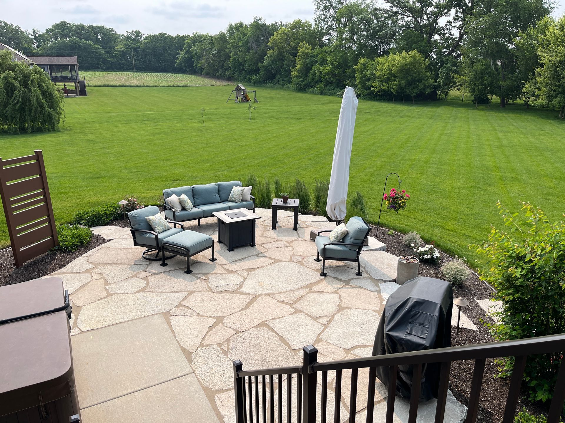 Patio with blue outdoor furniture, fire pit, and grill overlooking a large grassy yard and trees.