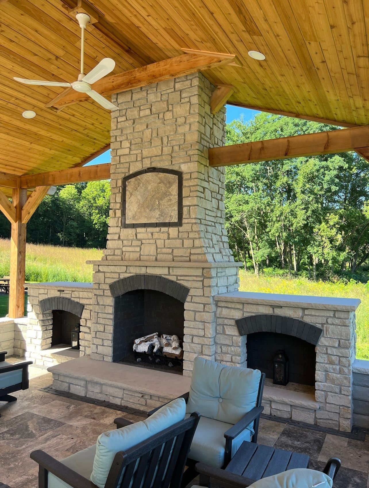 Outdoor stone fireplace with three openings, under a wooden roof, with seating.