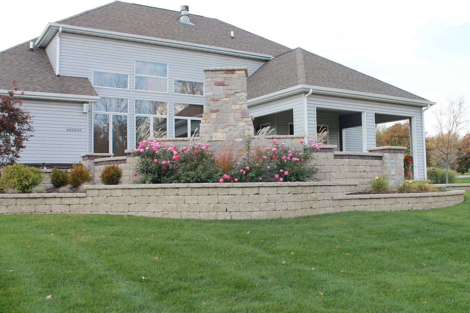 Beige brick retaining wall with landscaping and flowering bushes in front of a house.