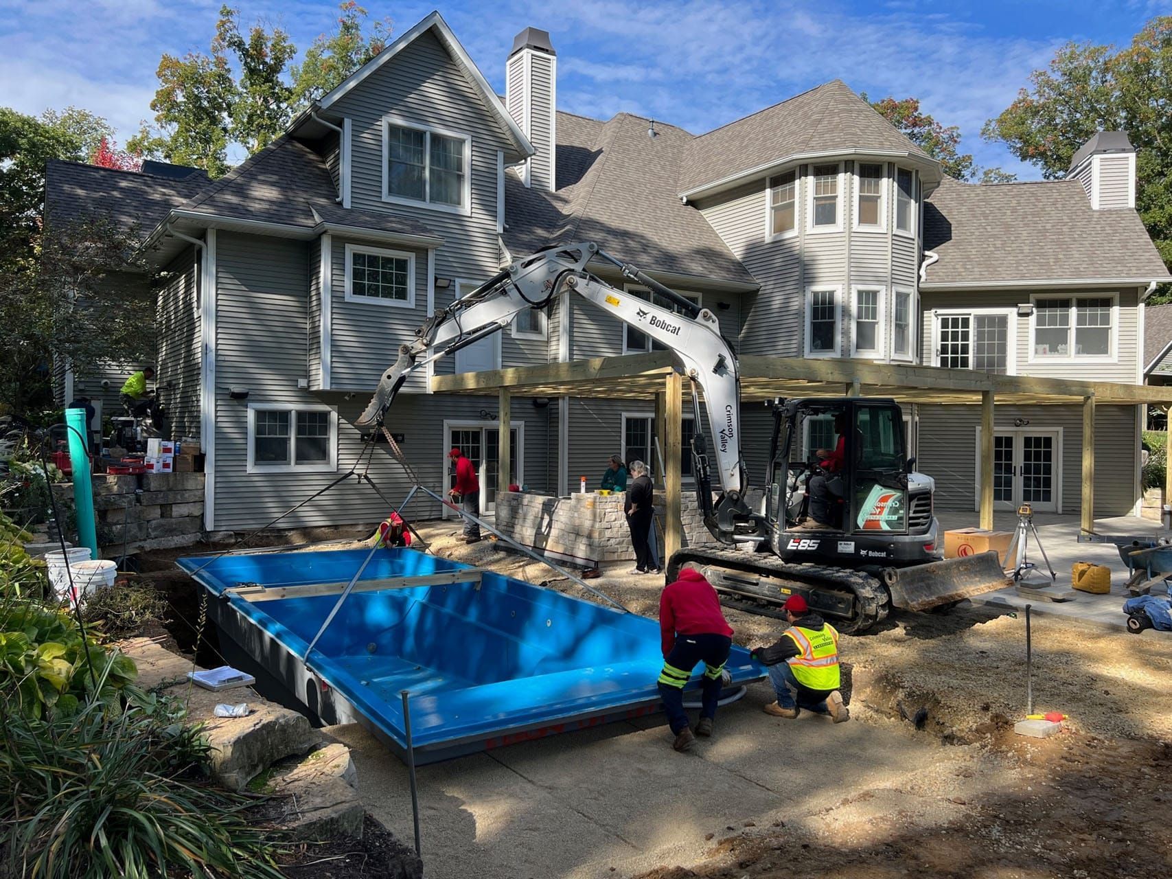 Pool installation underway behind a gray house. An excavator, workers, and a blue pool shell are visible.