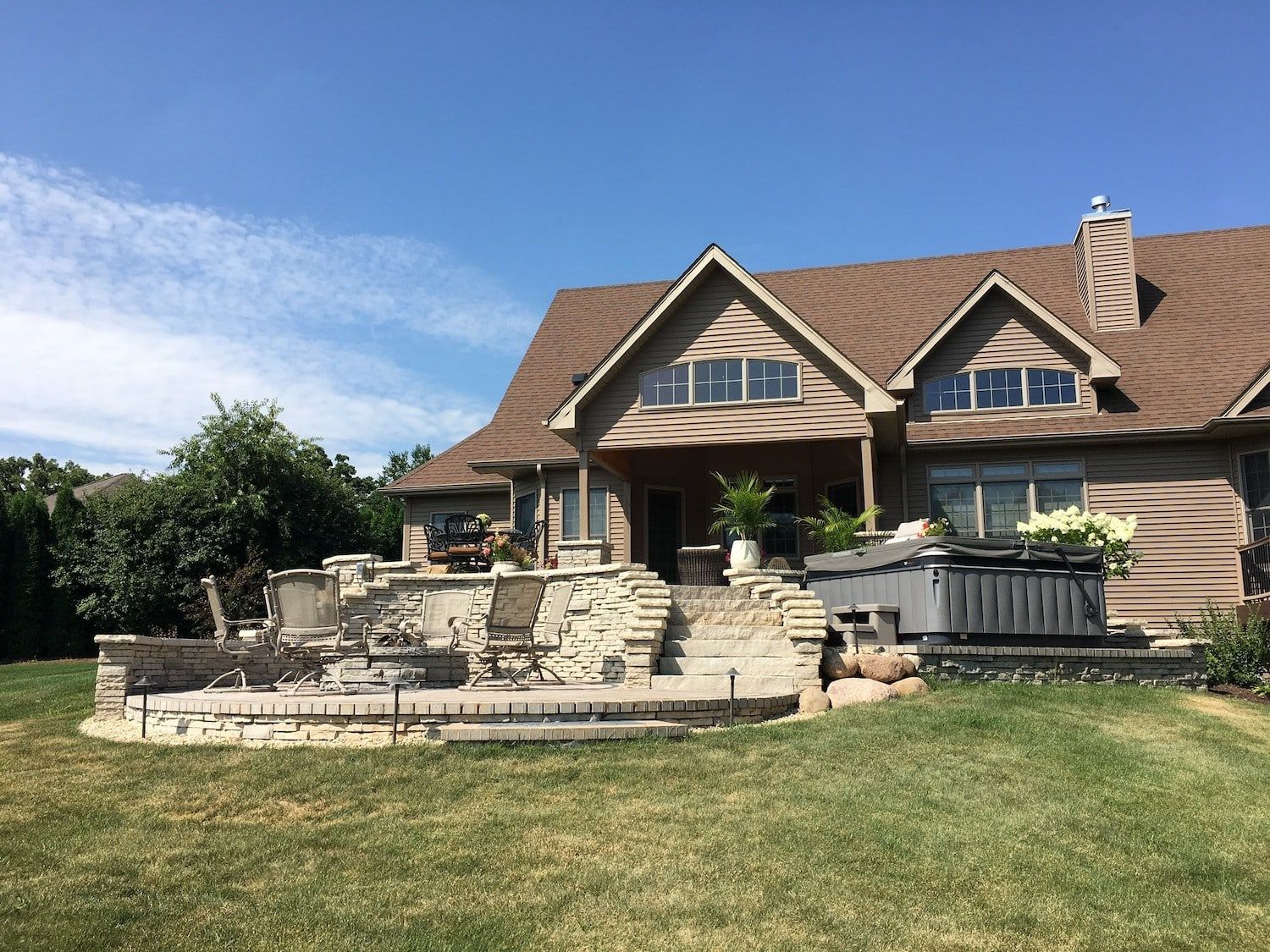 Large house with stone patio, hot tub, and green lawn under a blue sky.
