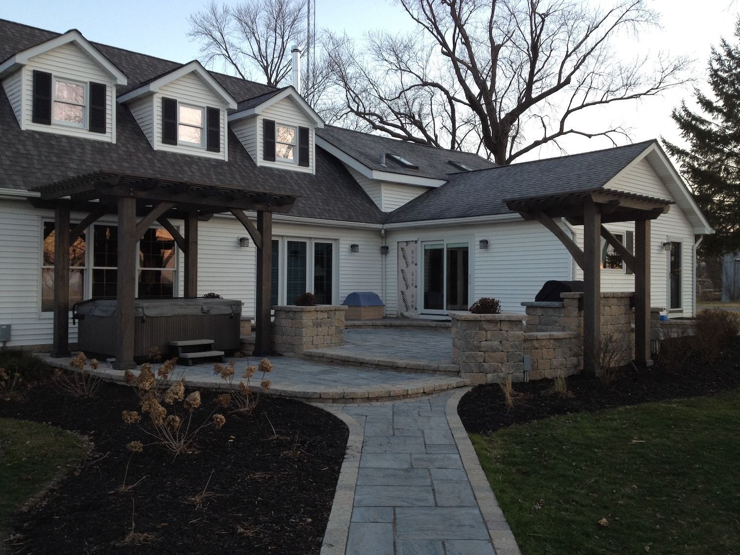 Backyard patio with a hot tub, pergola, and stone walkway leading to a house with dormer windows.