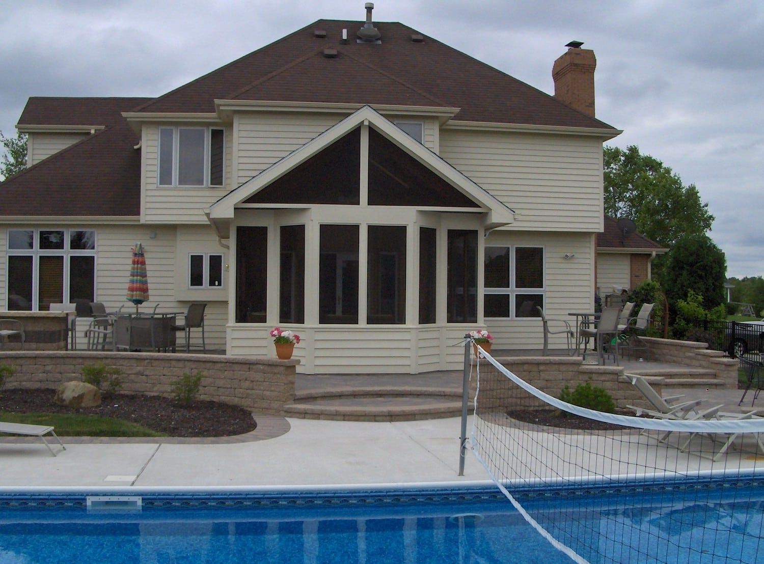 Back of beige house with attached screened porch, swimming pool, and volleyball net on a cloudy day.