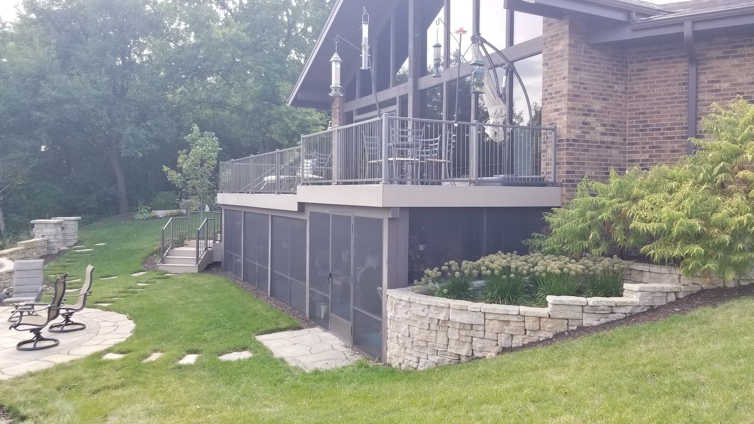House with deck, screened-in area, and stone retaining walls. Green grass and trees surround the structure.