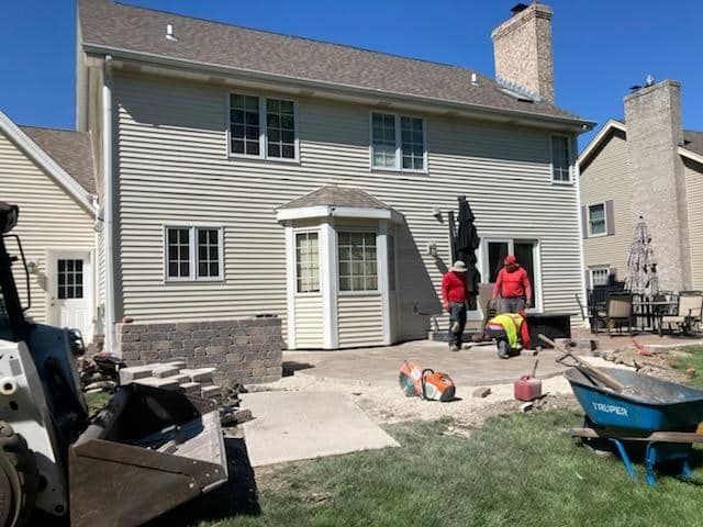 Backyard patio construction; workers near house with bay window and sliding door, machinery and materials visible.