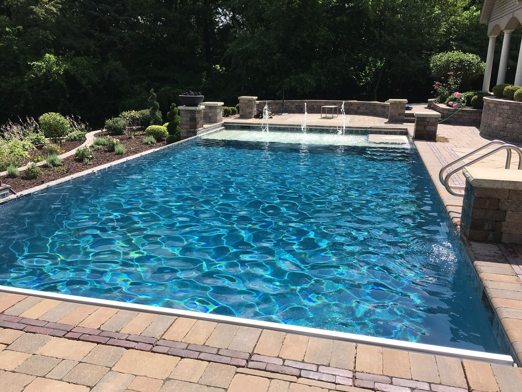 Rectangular outdoor swimming pool with blue water, surrounded by brick patio, lush greenery.