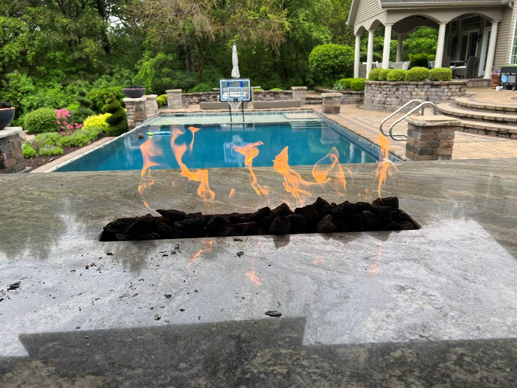 Fire pit flames on patio with a pool in the background. Lush greenery, blue water, and a home.