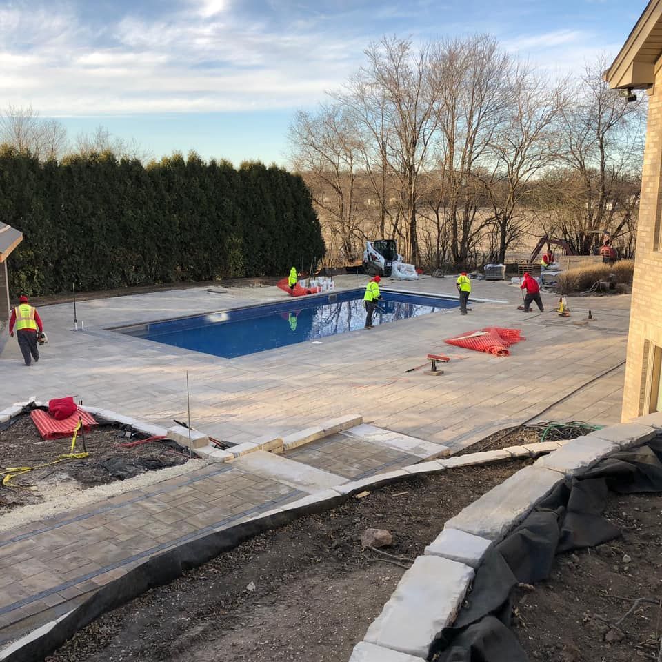 Construction workers around a rectangular pool, laying pavers. Bright sky, trees in the background.