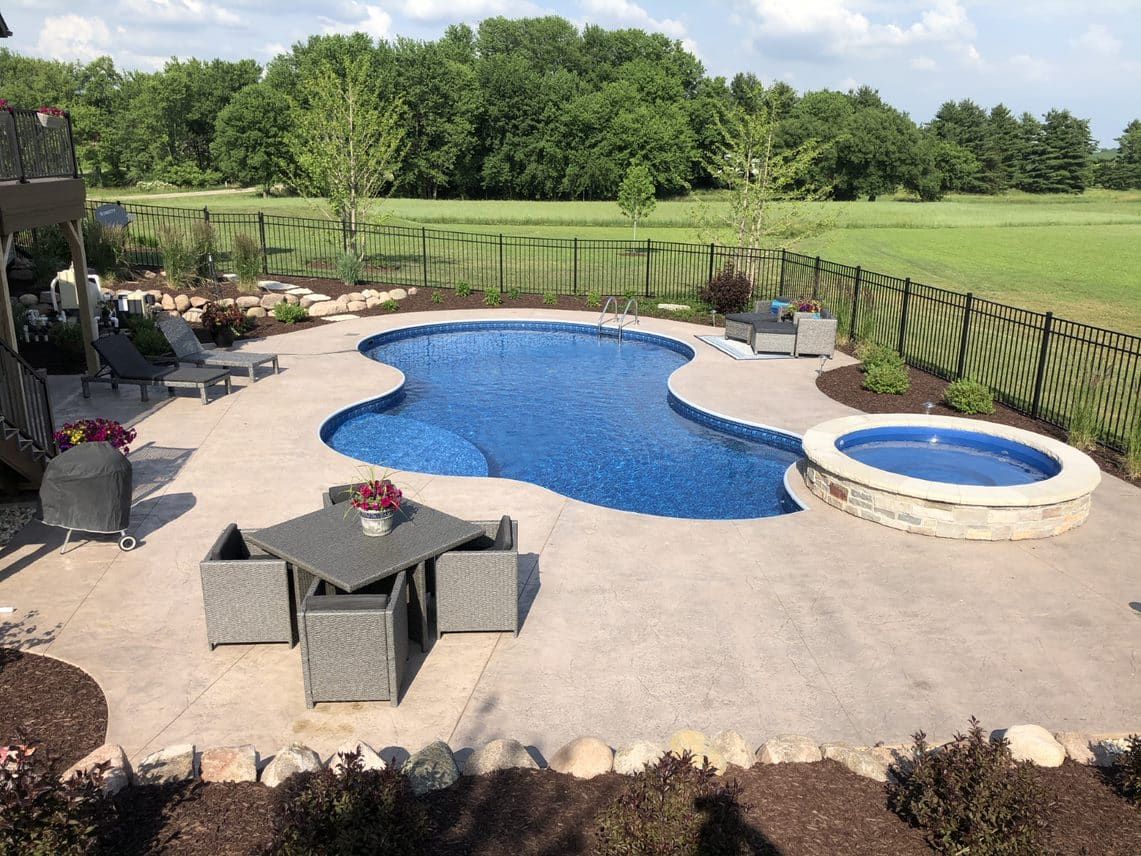 Backyard with a curvy blue pool, hot tub, patio furniture, and a fence. Trees and greenery in the background.