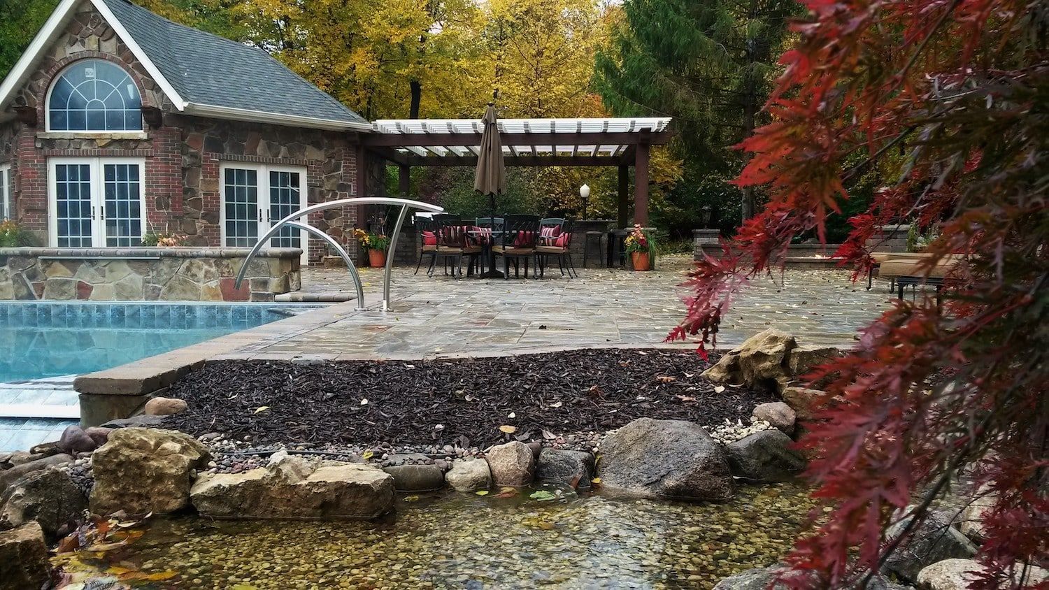 Stone house with pool, patio, and pergola surrounded by autumn foliage.