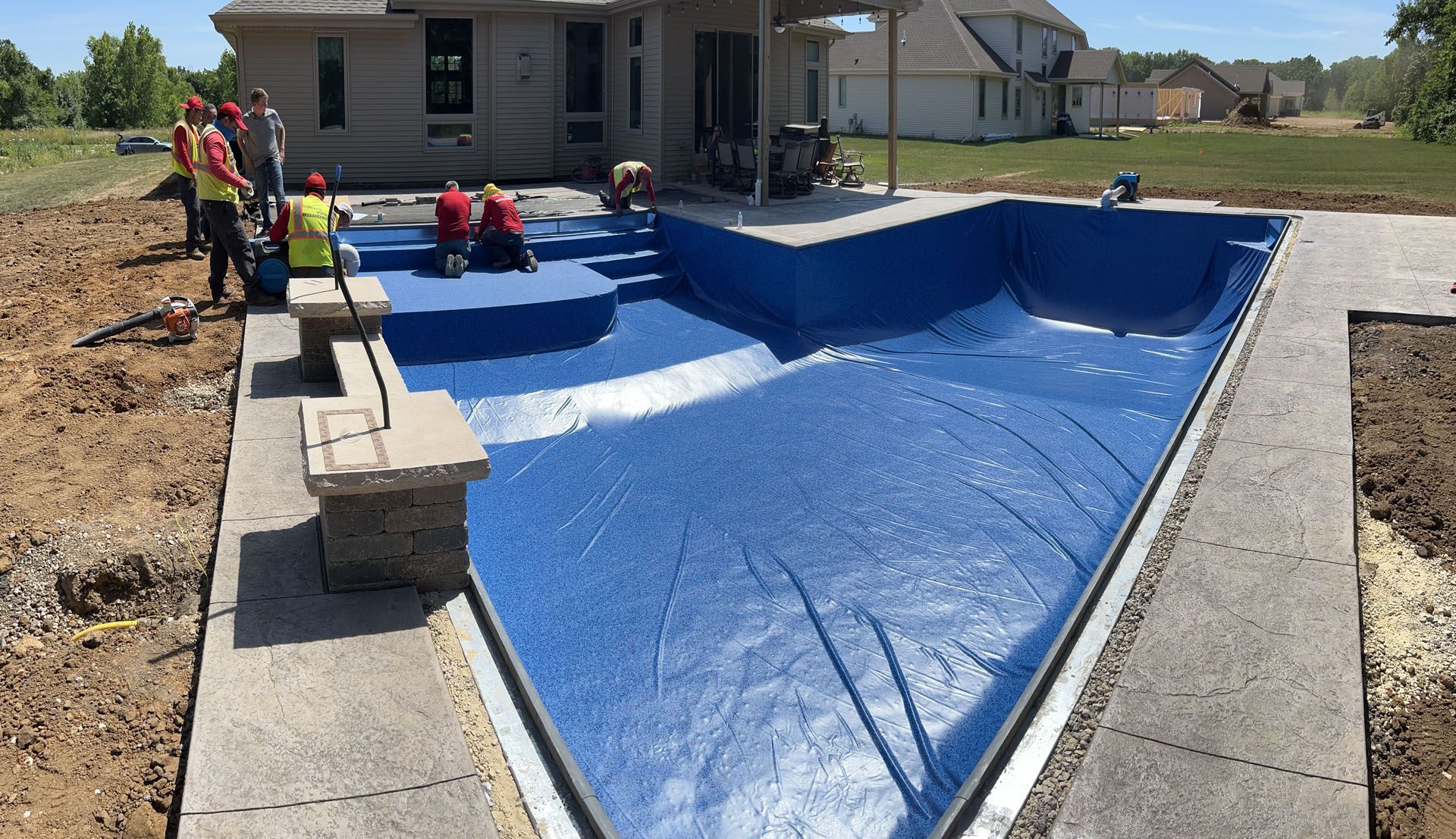 Workers installing a blue pool liner in an in-ground pool surrounded by concrete and landscaping.