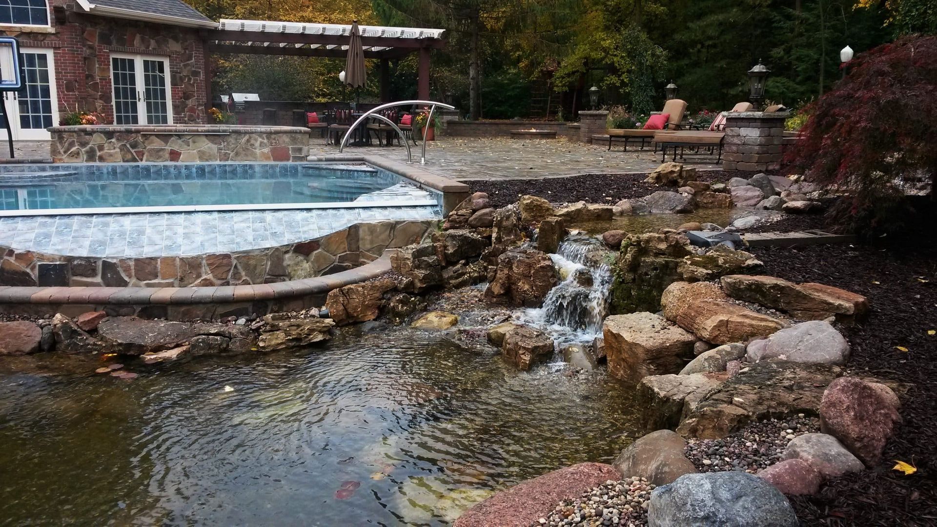 Backyard pool with waterfall feature flowing into a pond, adjacent to patio with seating.