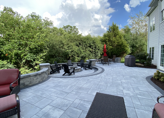 A backyard stone patio with a fire pit area, outdoor seating, and a red umbrella, surrounded by trees and a house.