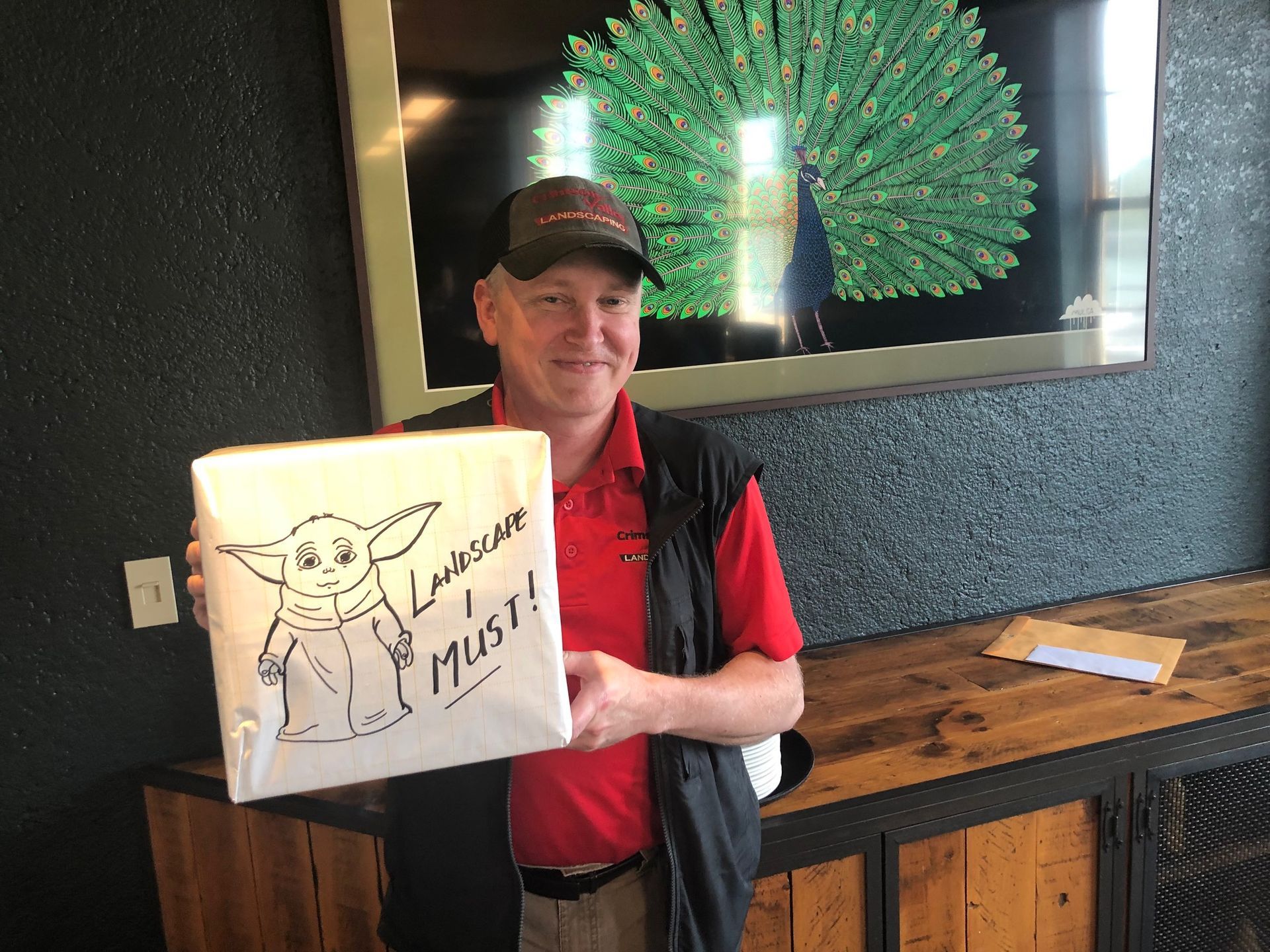A person in a red shirt and cap holds a gift box featuring a drawing of Baby Yoda.