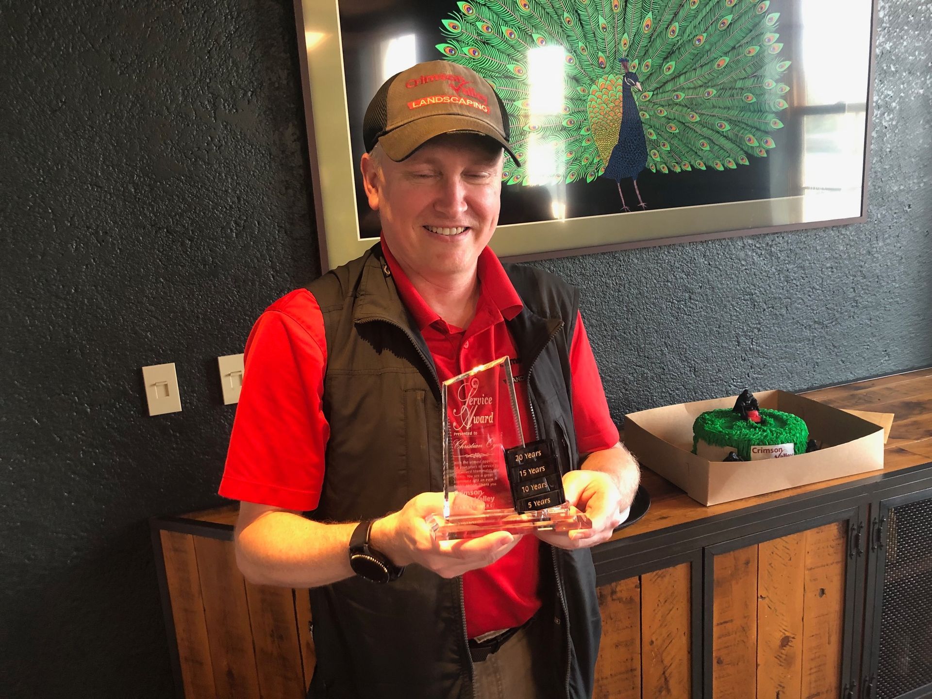 A person in a red polo, black vest, and cap smiles while holding a small, clear glass award in an indoor setting.