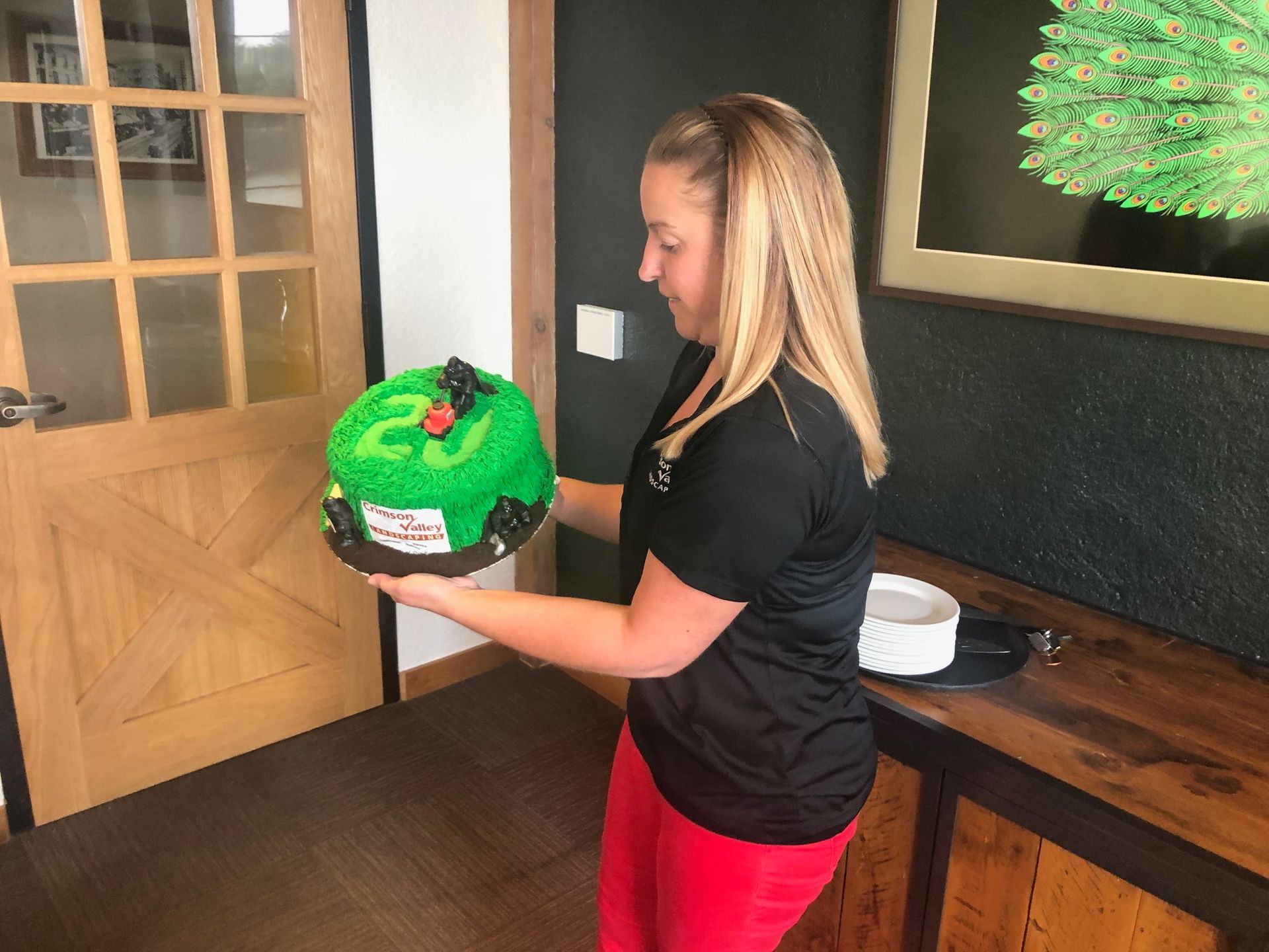 A person holds a bright green, round cake with a small black figure on top in an indoor room with wood-paneled walls.
