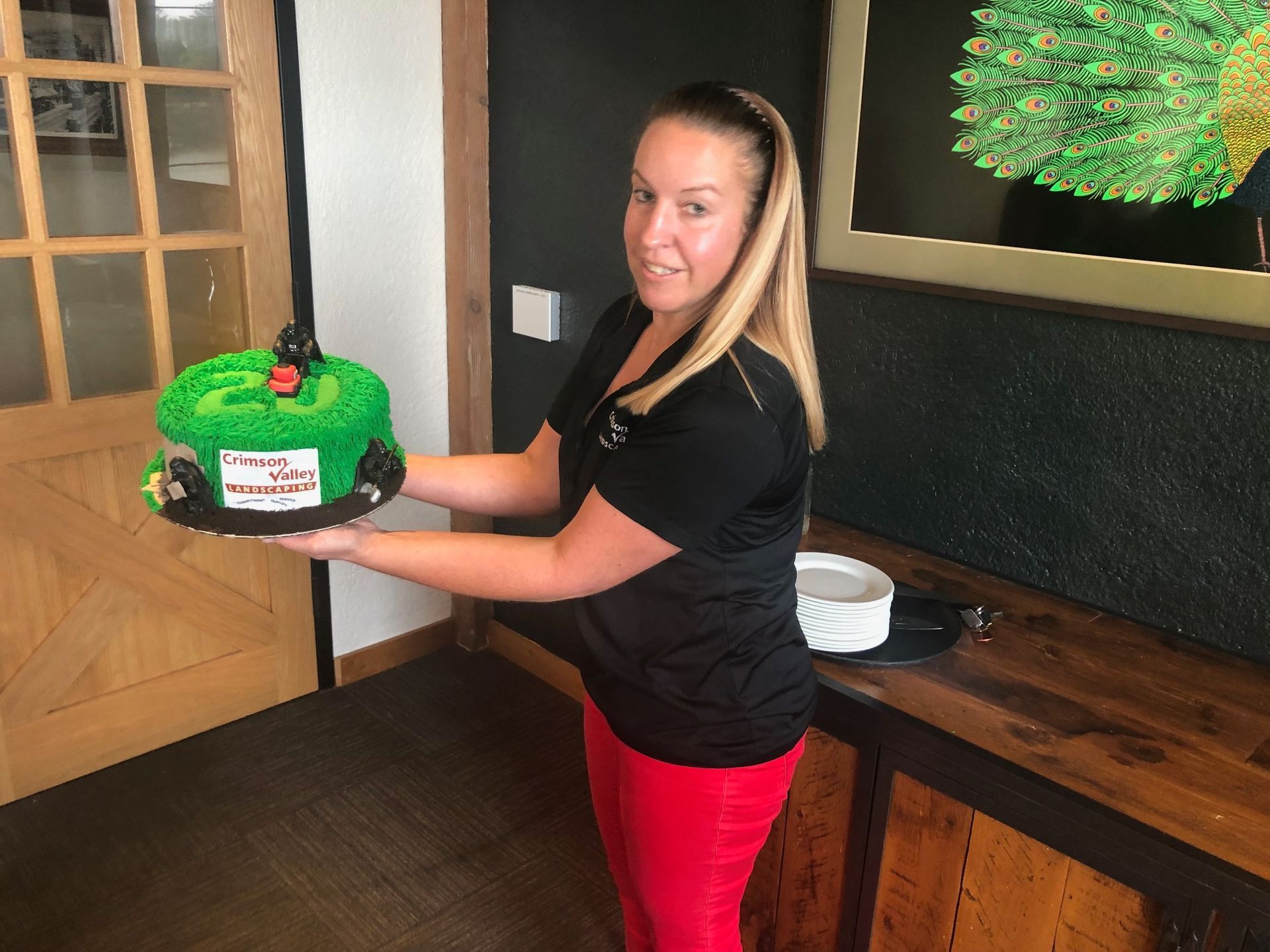 A person in a black shirt and red pants holds a round, green-frosted cake with small toy cars on top.