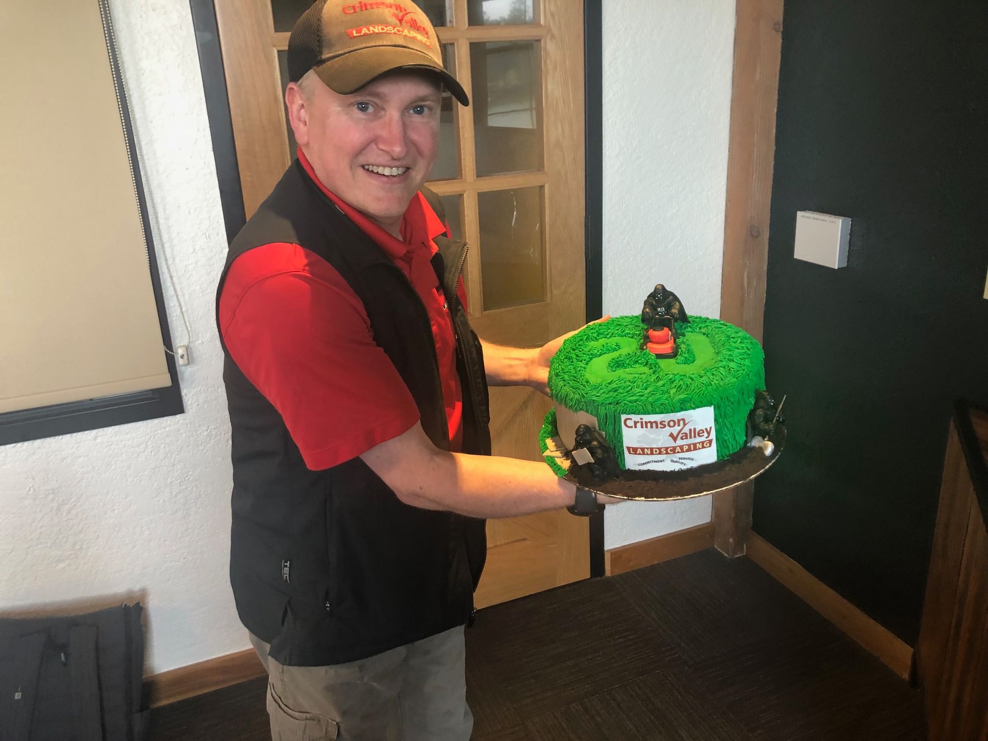 A person wearing a cap and red shirt holds a green cake topped with a black figurine in a brightly lit indoor space.
