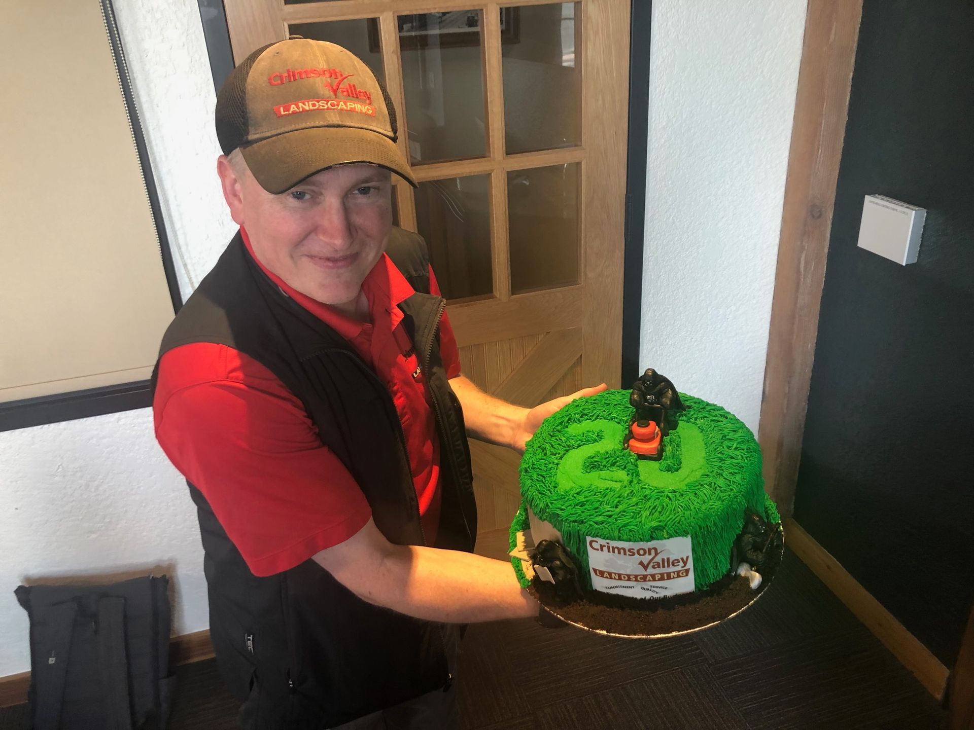 A person wearing a red shirt and cap holds a round green cake featuring the number 20 and a small toy tractor.