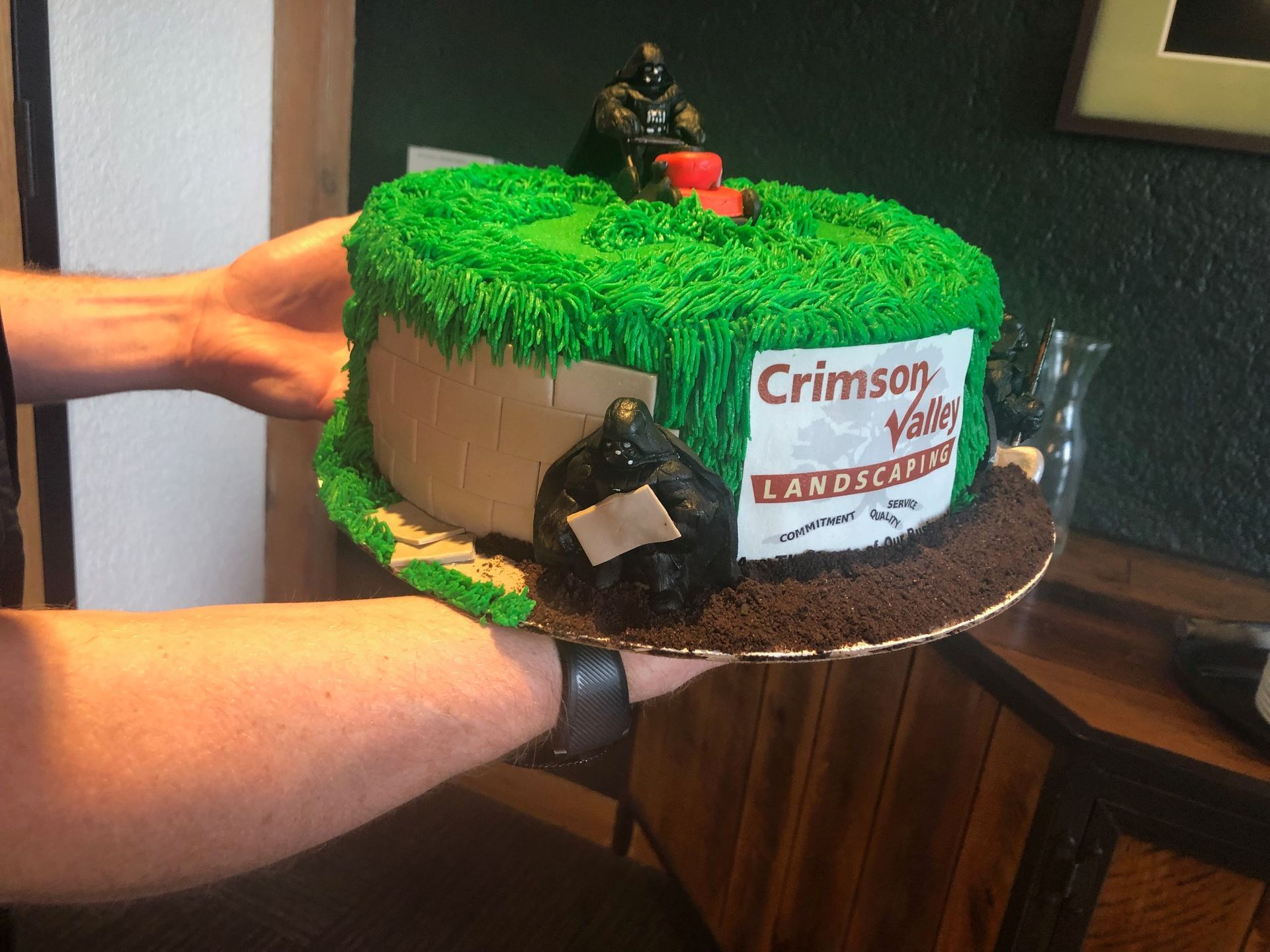 A person holds a green frosted cake decorated with a miniature lawnmower, a rock, and a Crimson Valley Landscaping logo.