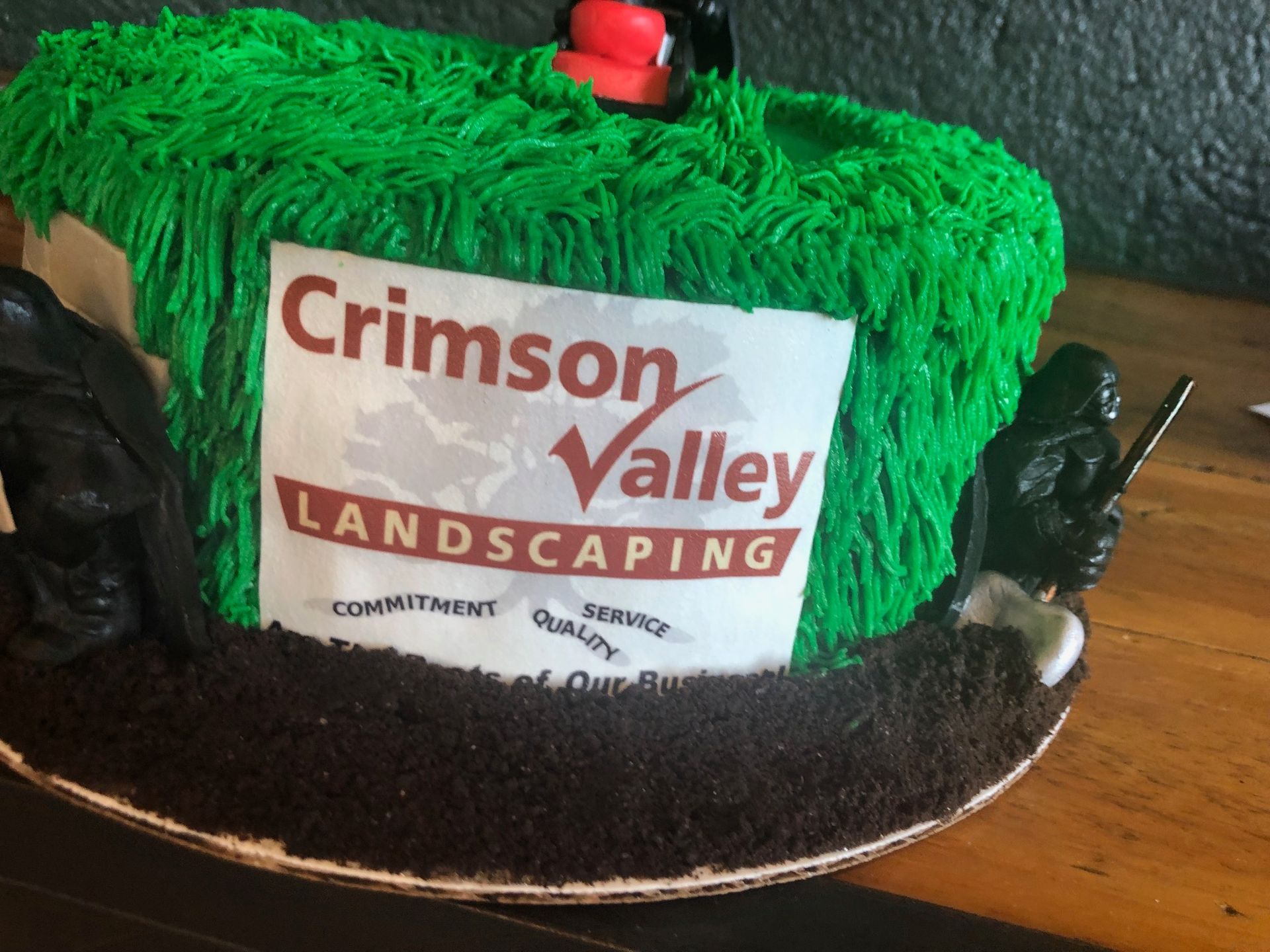 A cake decorated with green frosting as grass, a dirt-like crumb base, black figurines, and a Crimson Valley Landscaping logo.