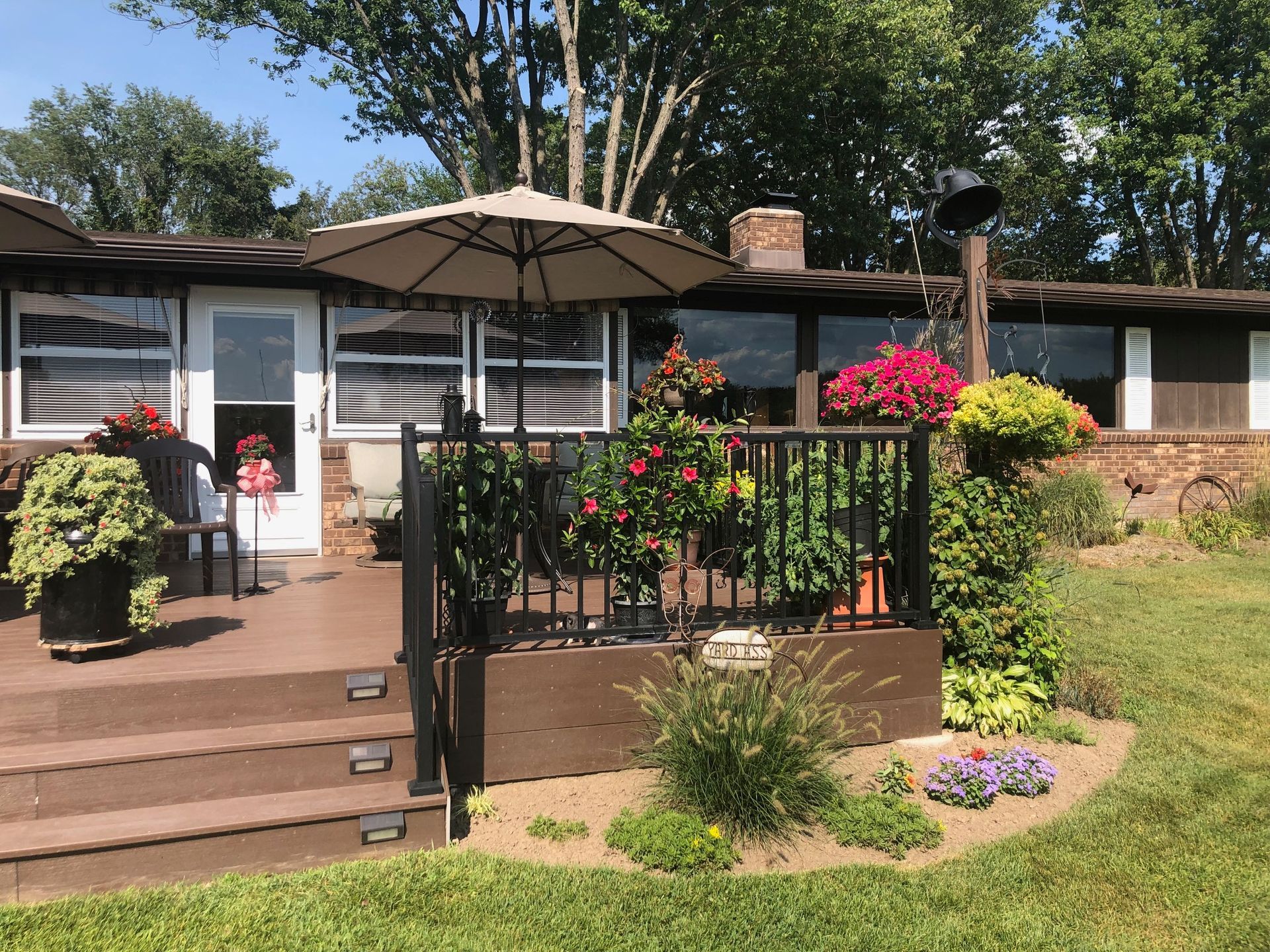 A sunny wooden deck with potted plants, hanging flower baskets, and an open patio umbrella in front of a house.