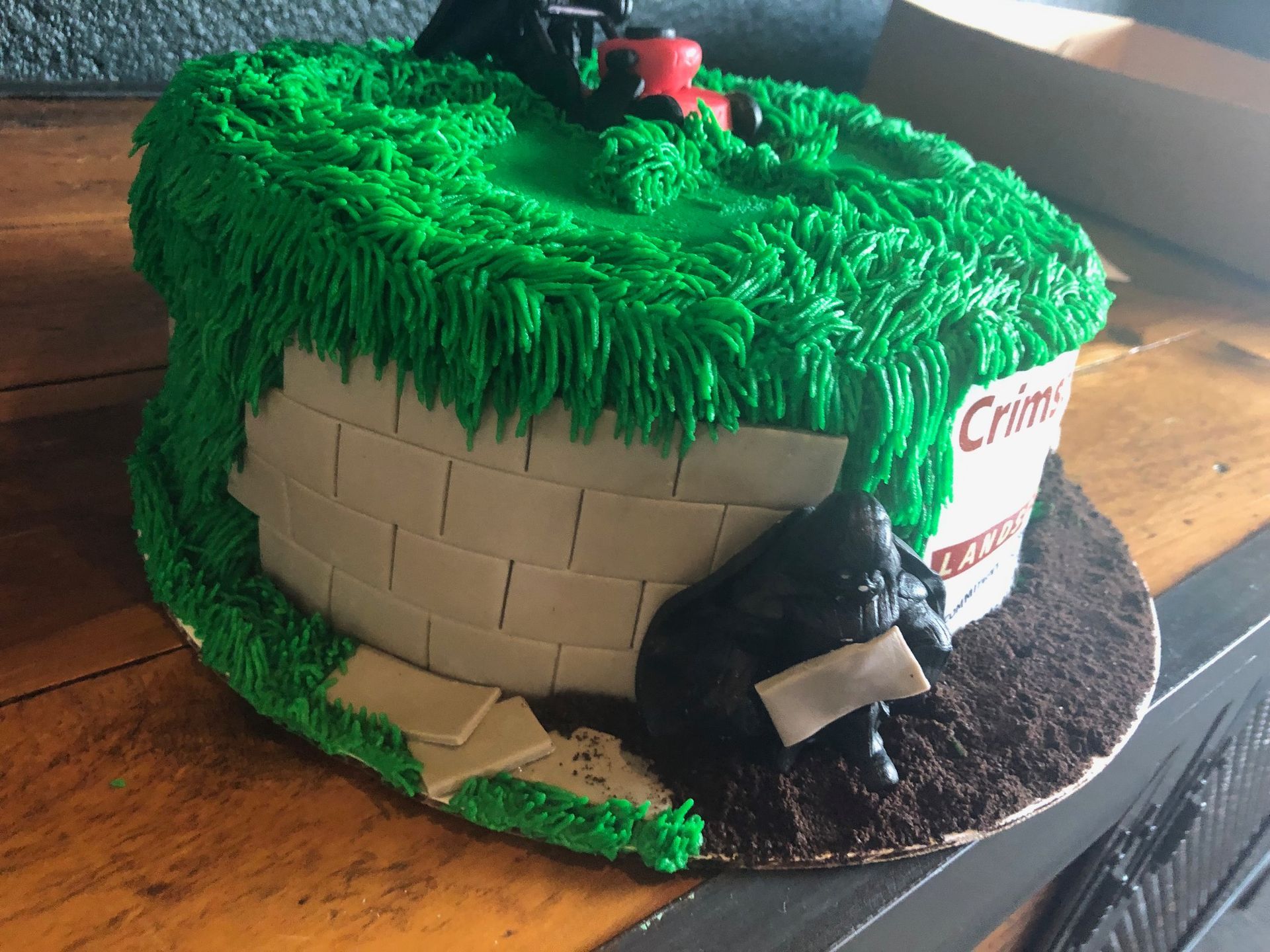 A round cake decorated like a grassy lawn with a brick wall, a lawnmower on top, and a figure sitting in the dirt.