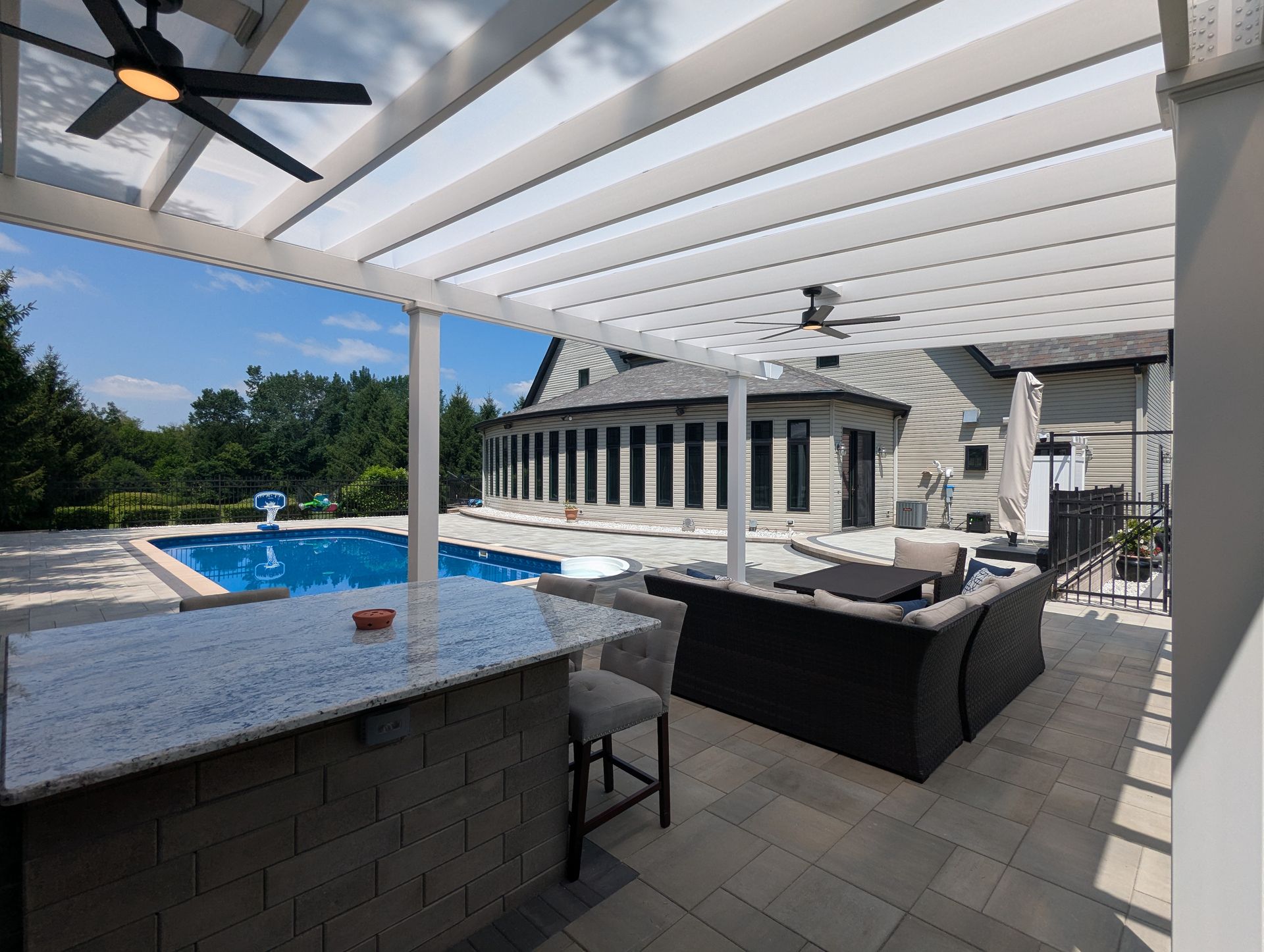 A shaded patio with a stone bar, grey stools, and outdoor seating overlooking a swimming pool and a house.