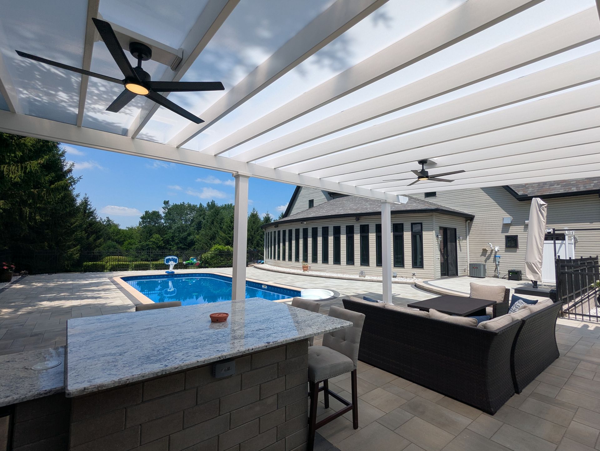 Outdoor patio with a granite-topped bar, wicker lounge seating, and ceiling fans under a white pergola, overlooking a pool.