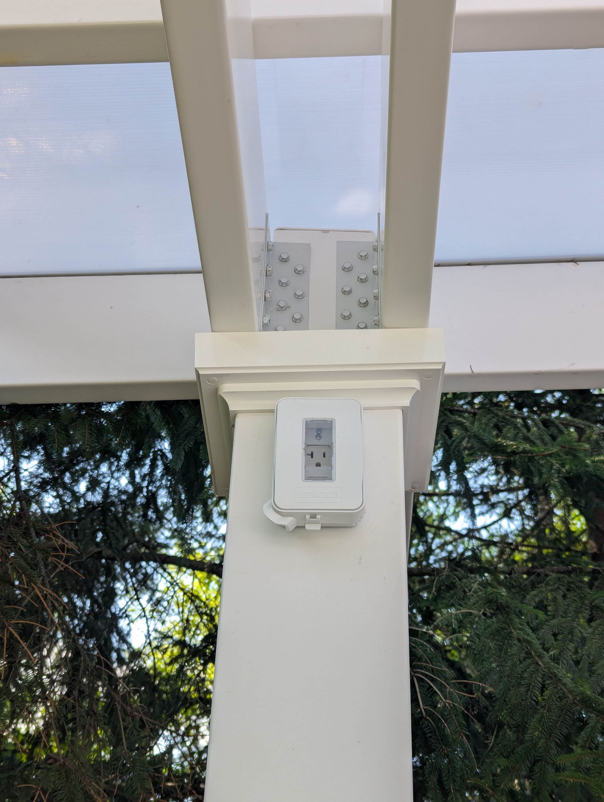 A white electrical outlet mounted on a pergola post, with metal brackets joining beams above.