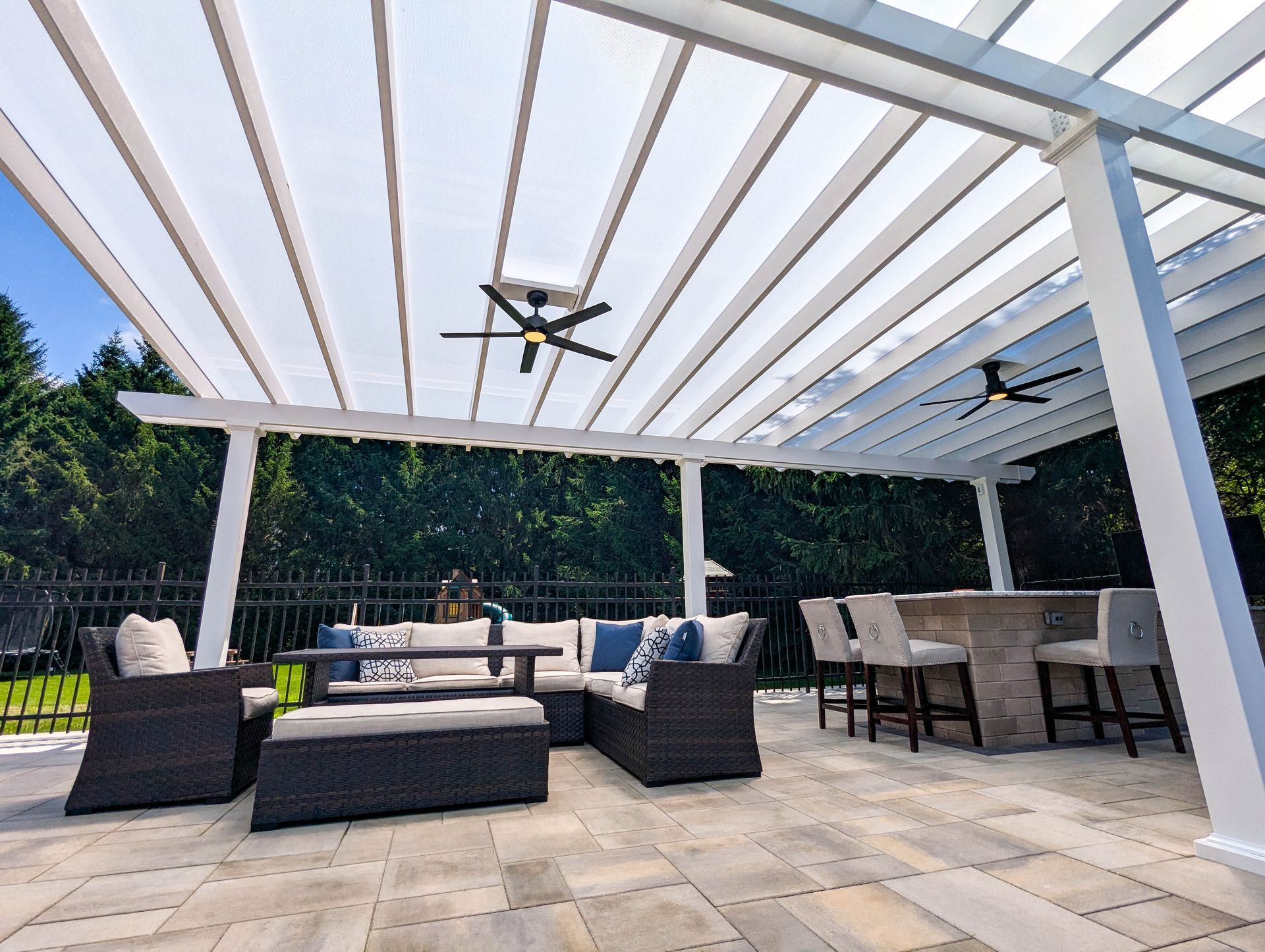 A white pergola covers a stone patio with dark wicker outdoor seating, a dining set, and two ceiling fans.