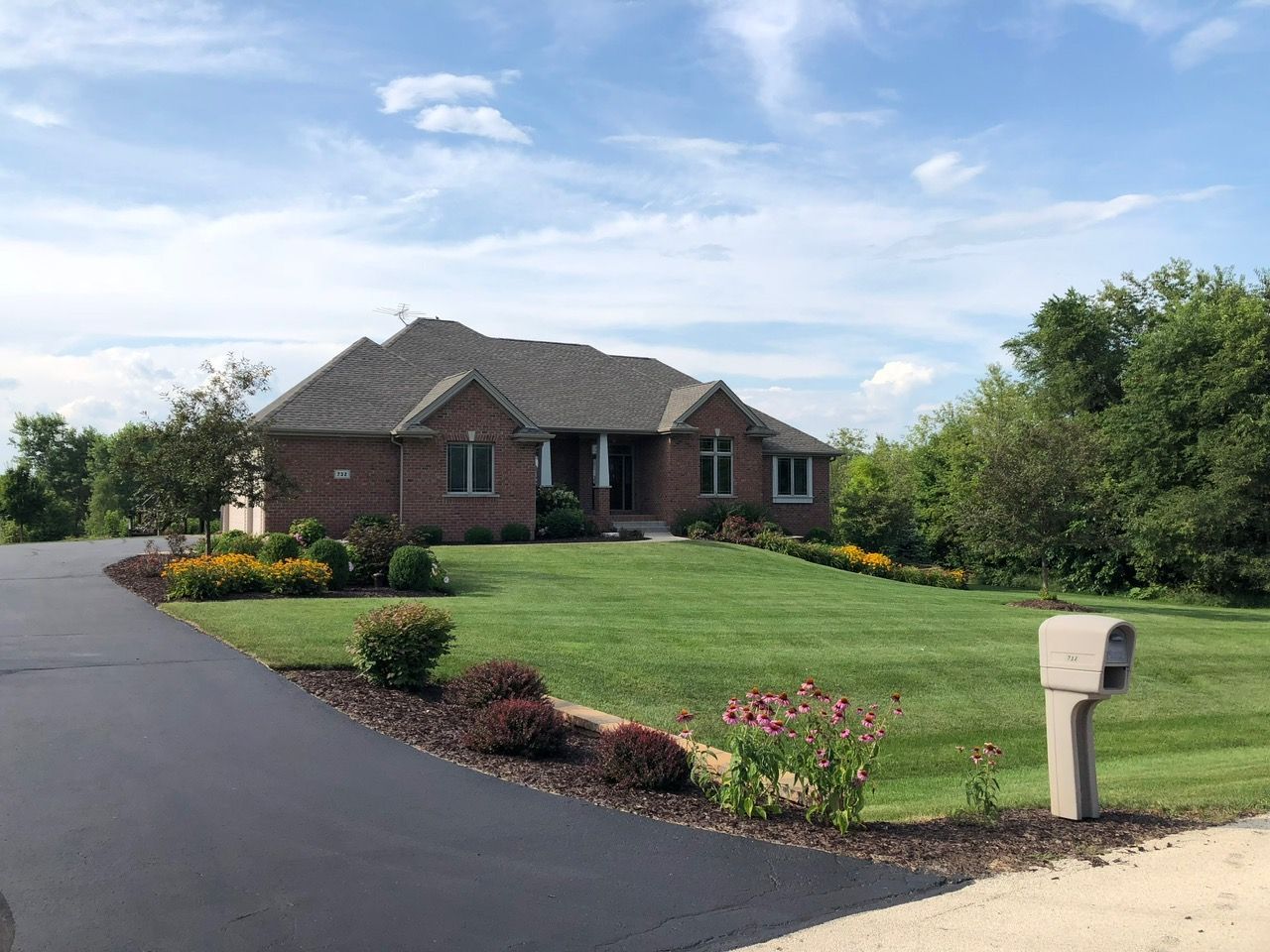 A brick house with a dark shingled roof, a green lawn, and a black asphalt driveway under a blue, cloudy sky.