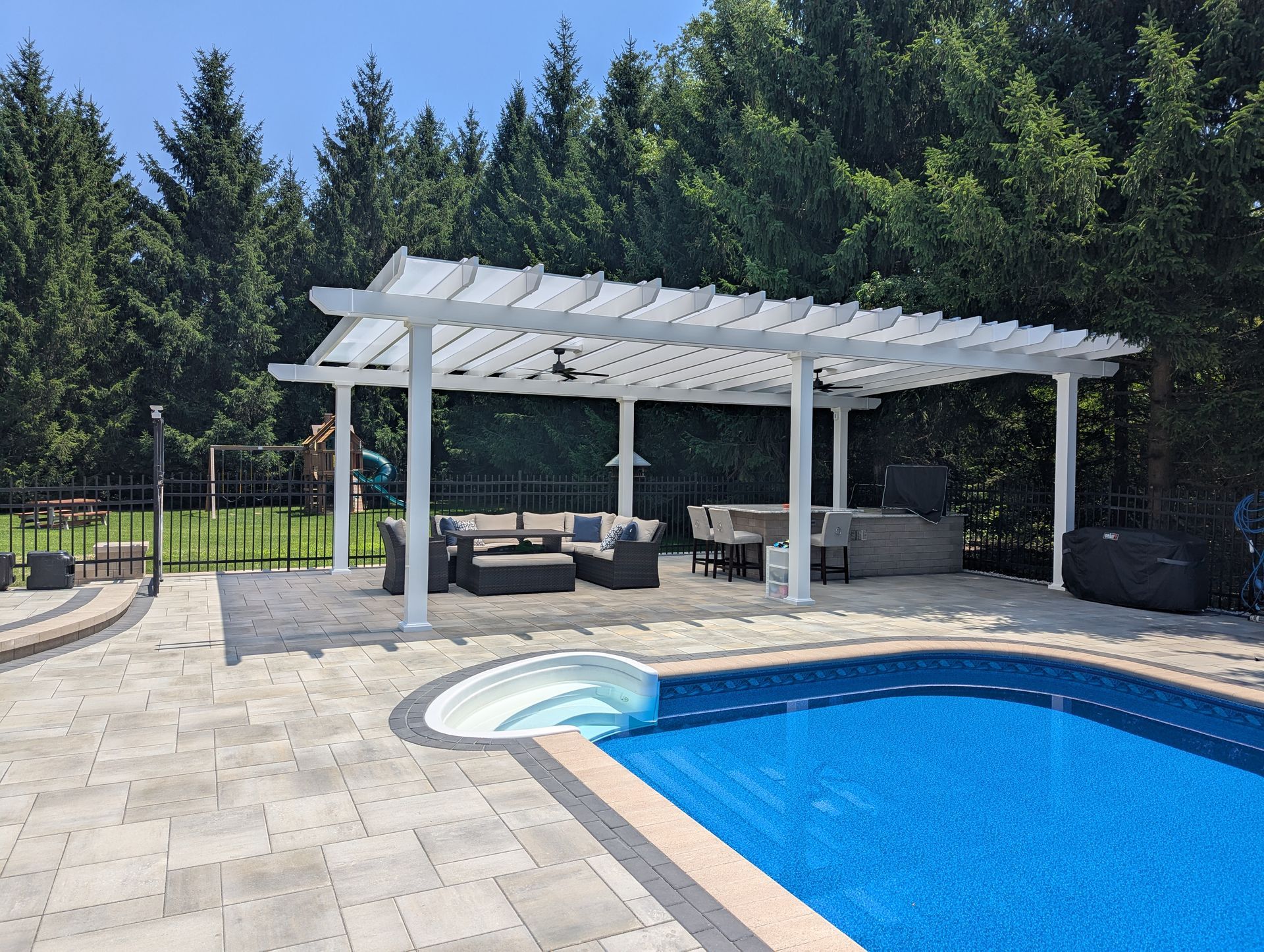 A white pergola over outdoor furniture on a stone patio next to a backyard swimming pool, surrounded by tall pine trees.