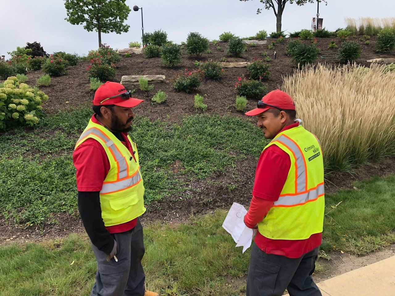 Two workers in red shirts and yellow safety vests stand outdoors discussing a document near a landscaped slope.