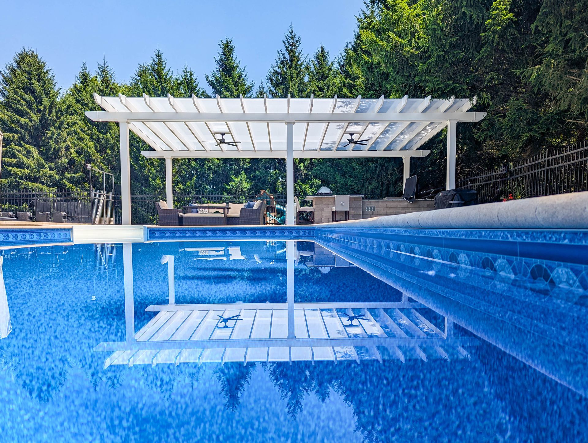 A white wooden pergola reflected in the blue water of a backyard swimming pool surrounded by evergreen trees.
